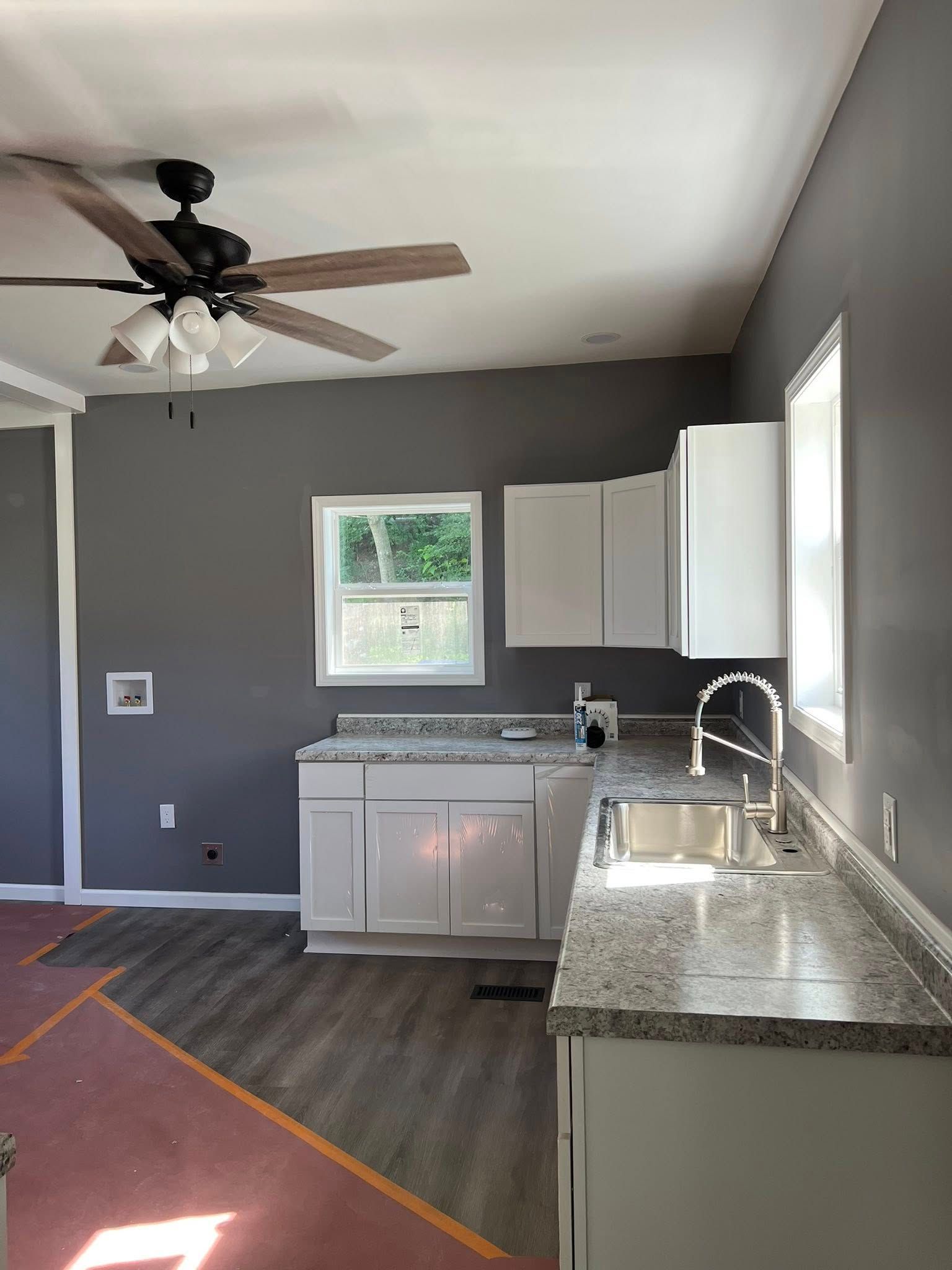 Empty kitchen with gray walls, white cabinets, granite counters, window, ceiling fan, and dark wood floor.