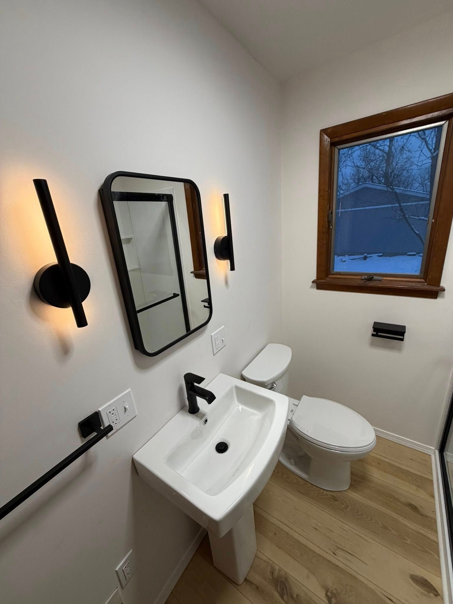 Small bathroom with white sink, toilet, wall mirror, black sconces, and a window with snowy view.