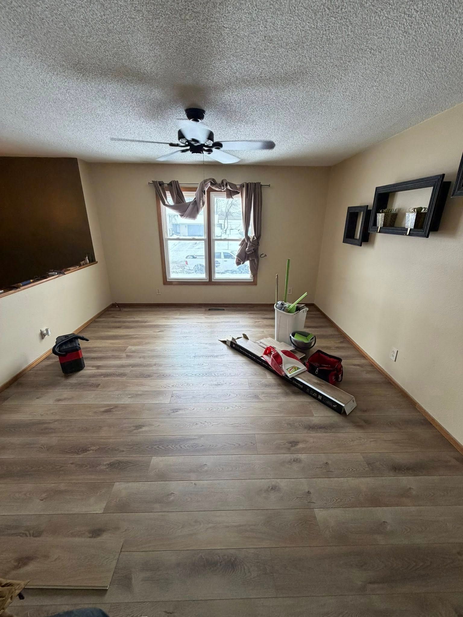 Empty beige room with wood-look floors, window, ceiling fan, and a red-and-white centerpiece on the floor