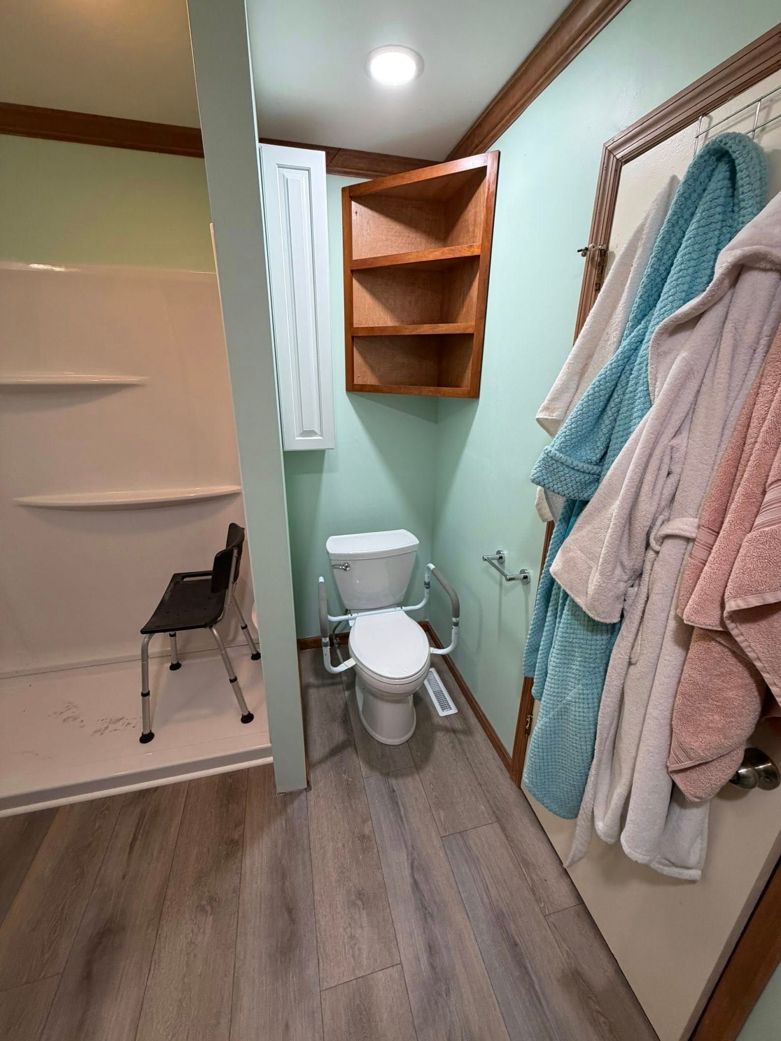 Small bathroom with toilet, corner shelves, towel rack, and striped bath mat on wood-look floor.
