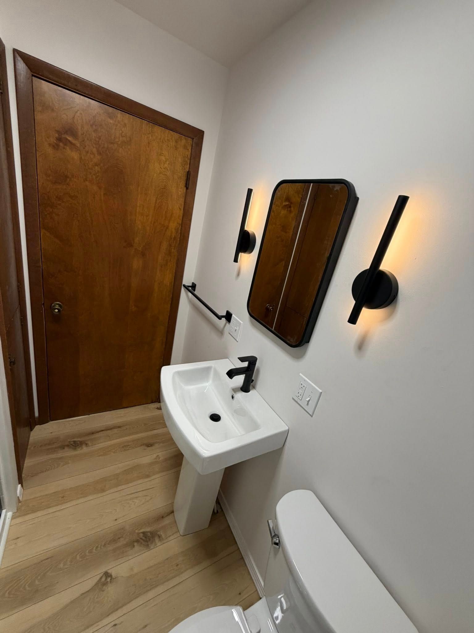 Small bathroom with wood door, pedestal sink, toilet, mirror, and wall sconces on light wood floor.