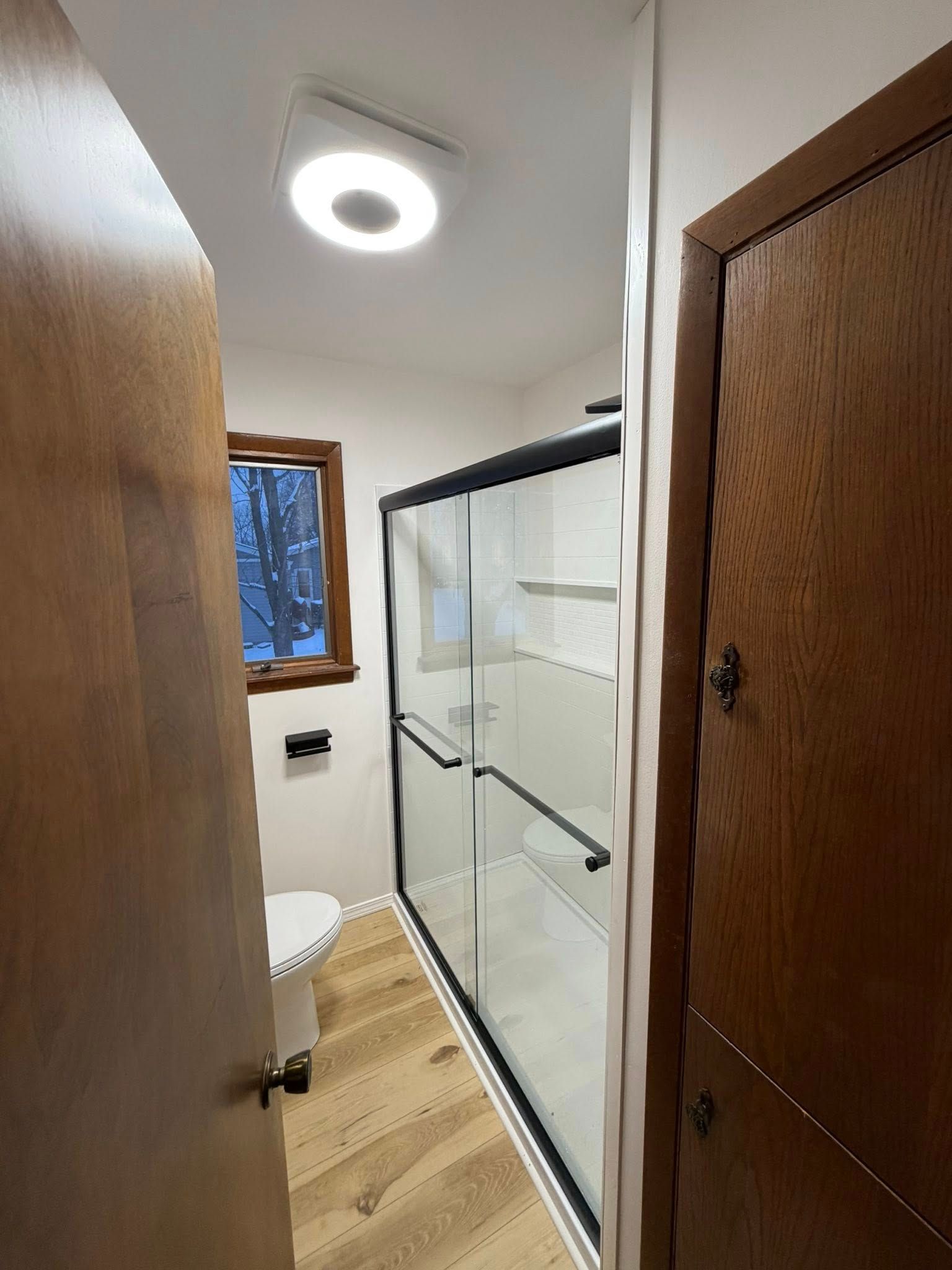 Narrow bathroom hallway with wood doors, glass shower, window, and circular ceiling light.