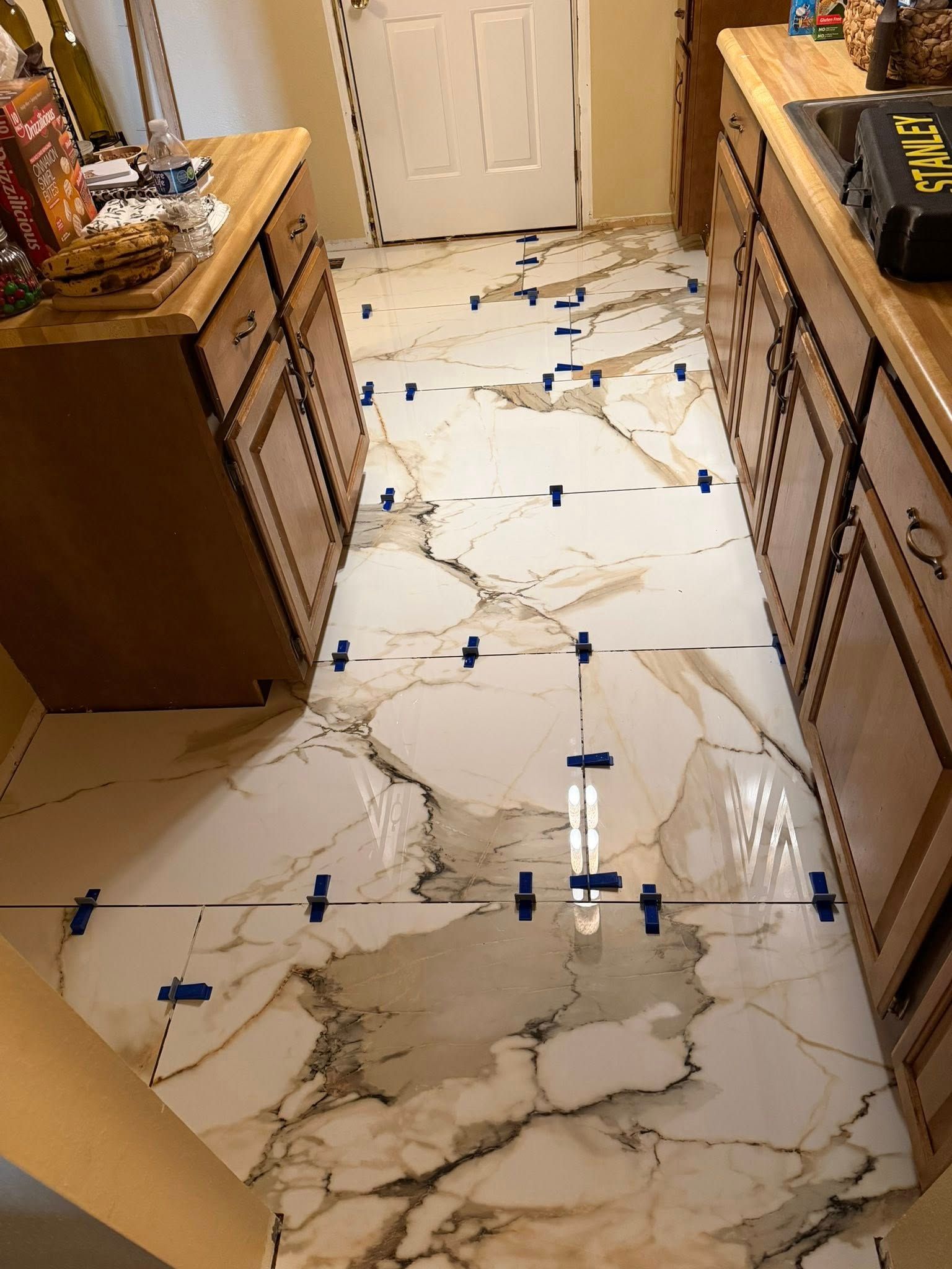 A tiled kitchen floor with blue tape markers between two wood cabinets and a white door.