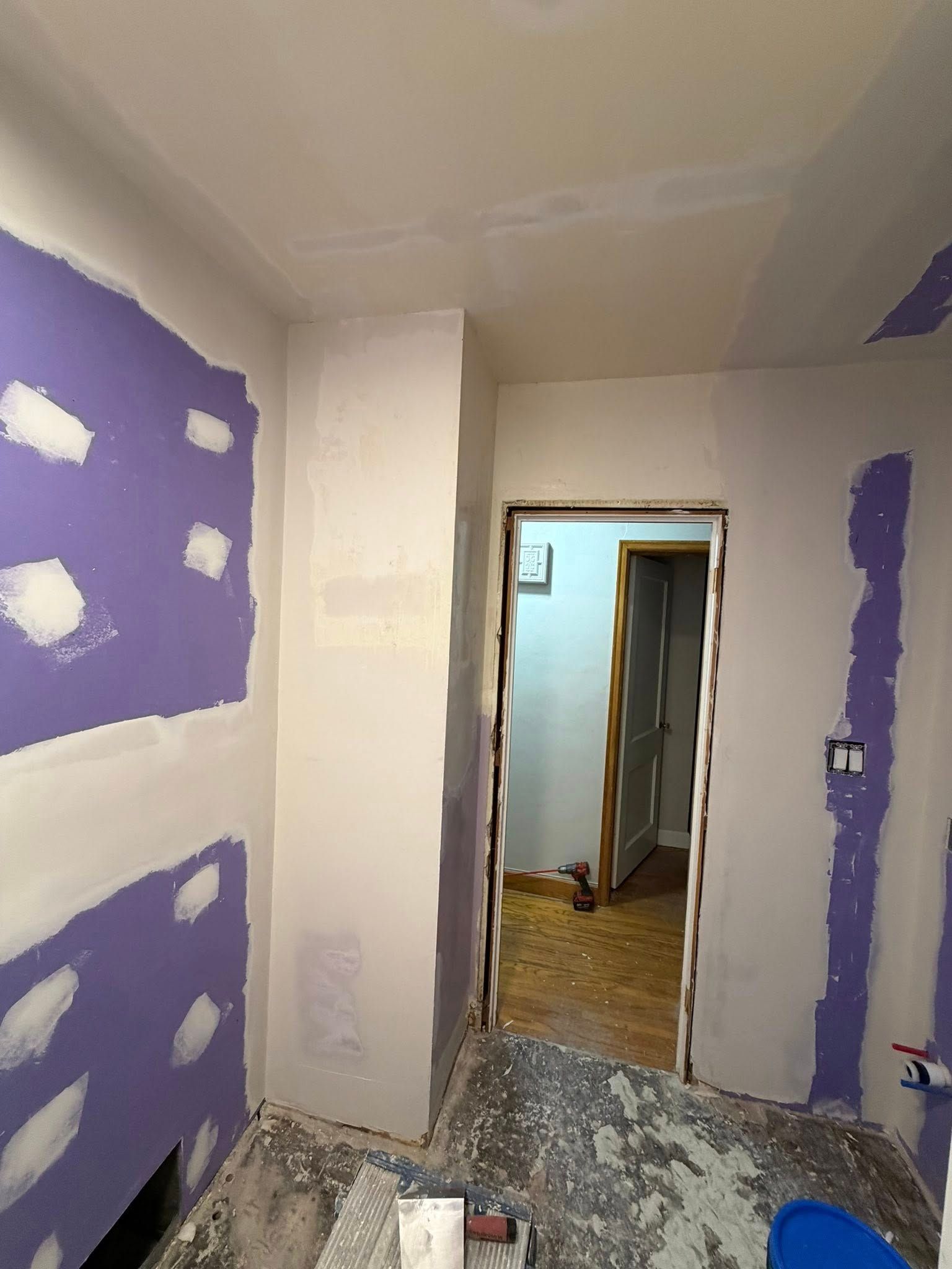 Narrow hallway under renovation with purple drywall patches and unfinished walls leading to a room beyond