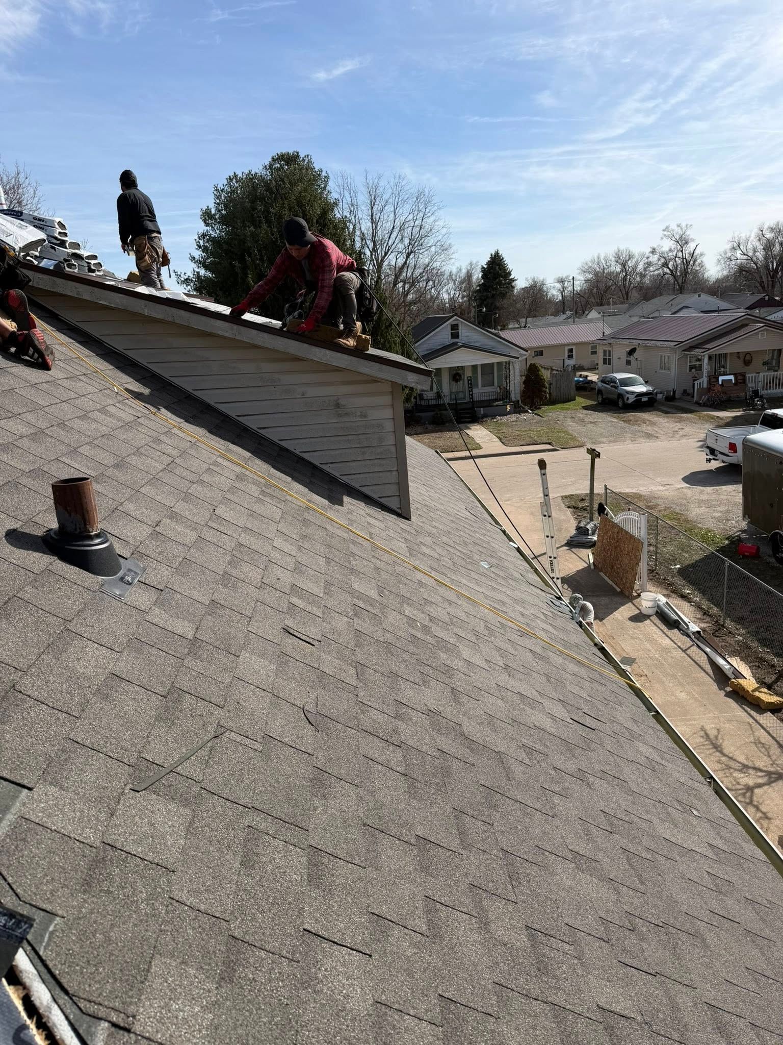 Roofers working on a shingled roof with a neighborhood in the background