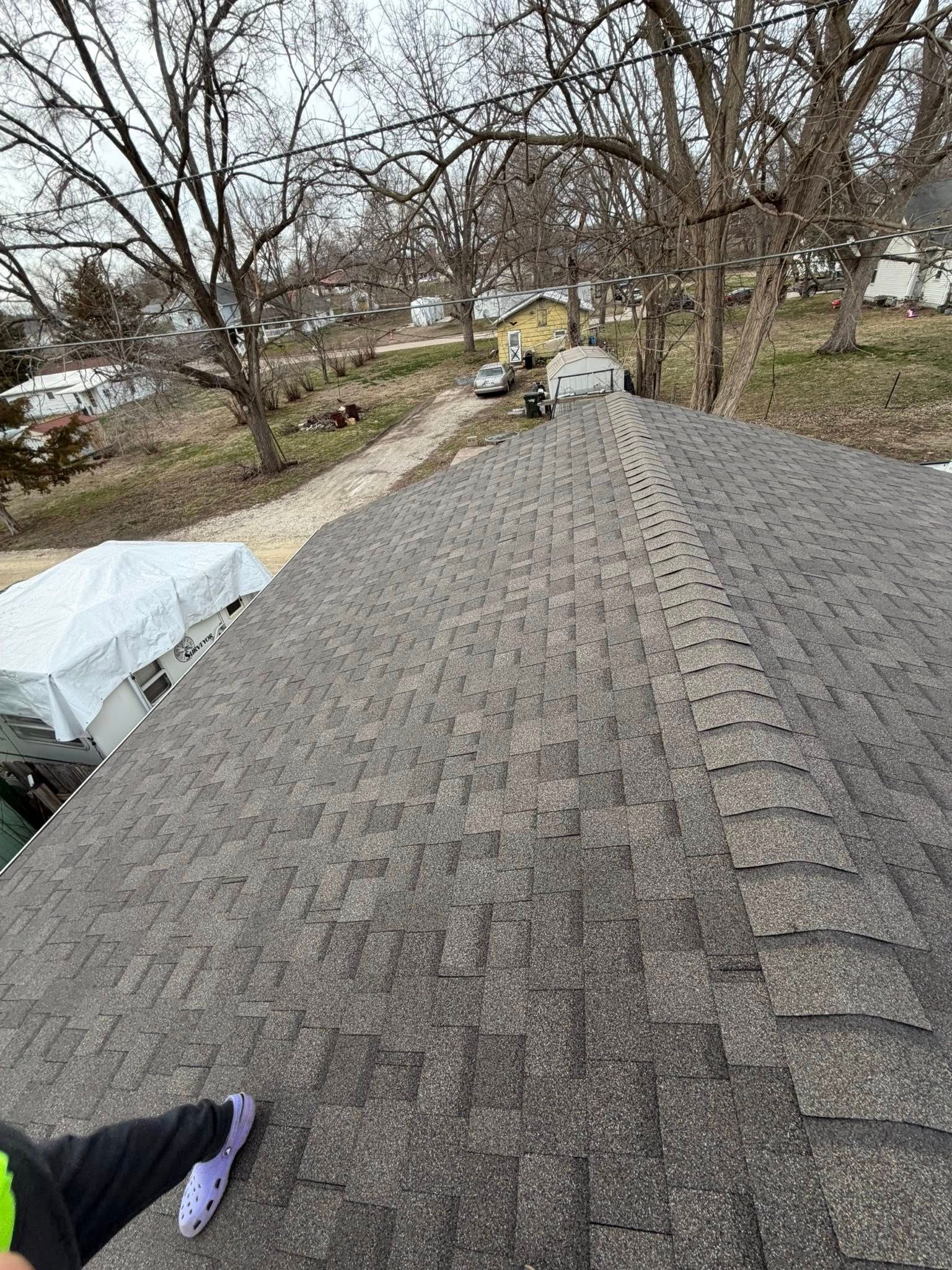 Roof shingles with workers in the distance and a gloved hand at the edge of the roof under bare trees