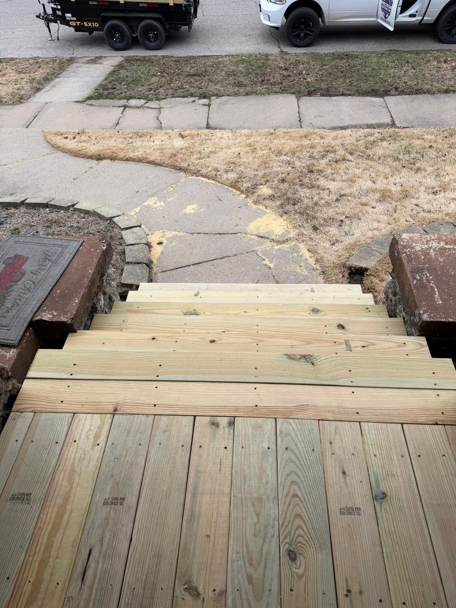 Wooden ramp leading from a porch to a sidewalk, with parked vehicles and a patchy lawn beyond.