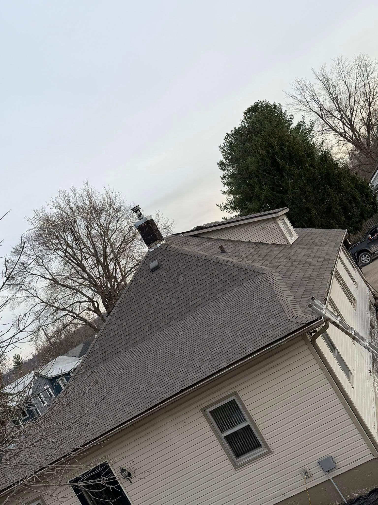 Tilted view of a beige house with gray roof and trees under an overcast sky