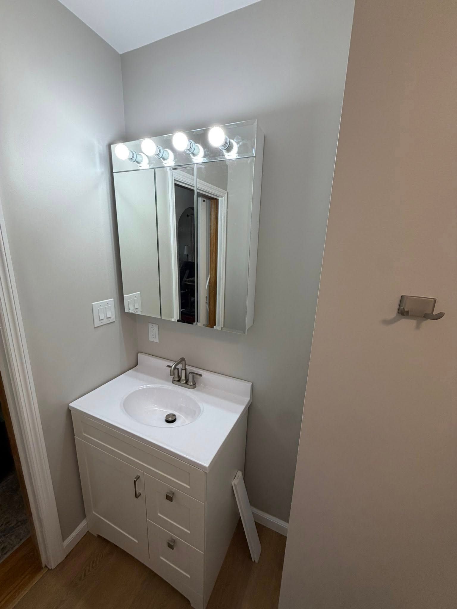 Small bathroom vanity with mirror and lit bulbs, white sink and cabinet in a narrow corner.