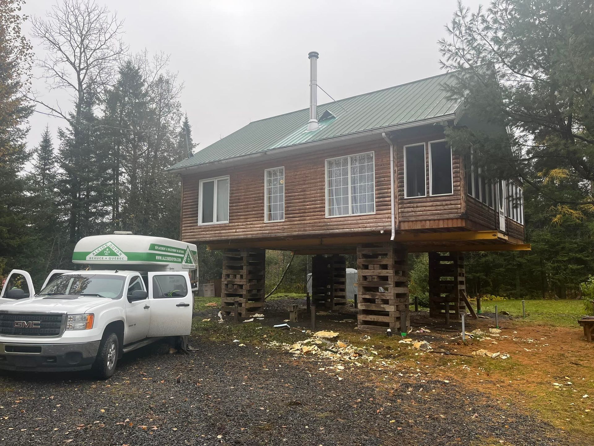 Une maison en bois sur pilotis de deux étages avec un toit vert, à côté d'un camion blanc à porte ouverte.