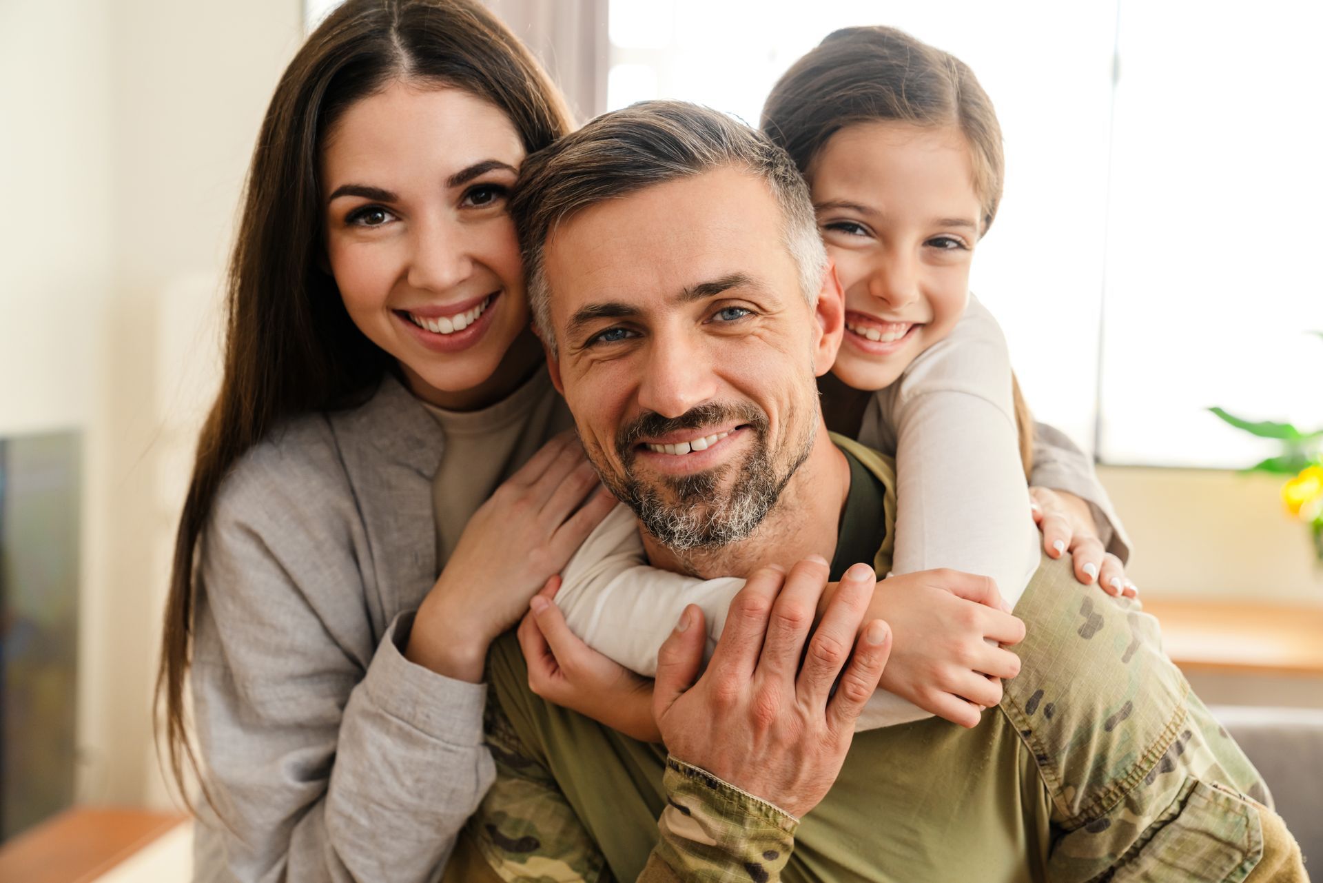 Smiling family of three hugging indoors, with a child resting on a man’s back and a woman beside them