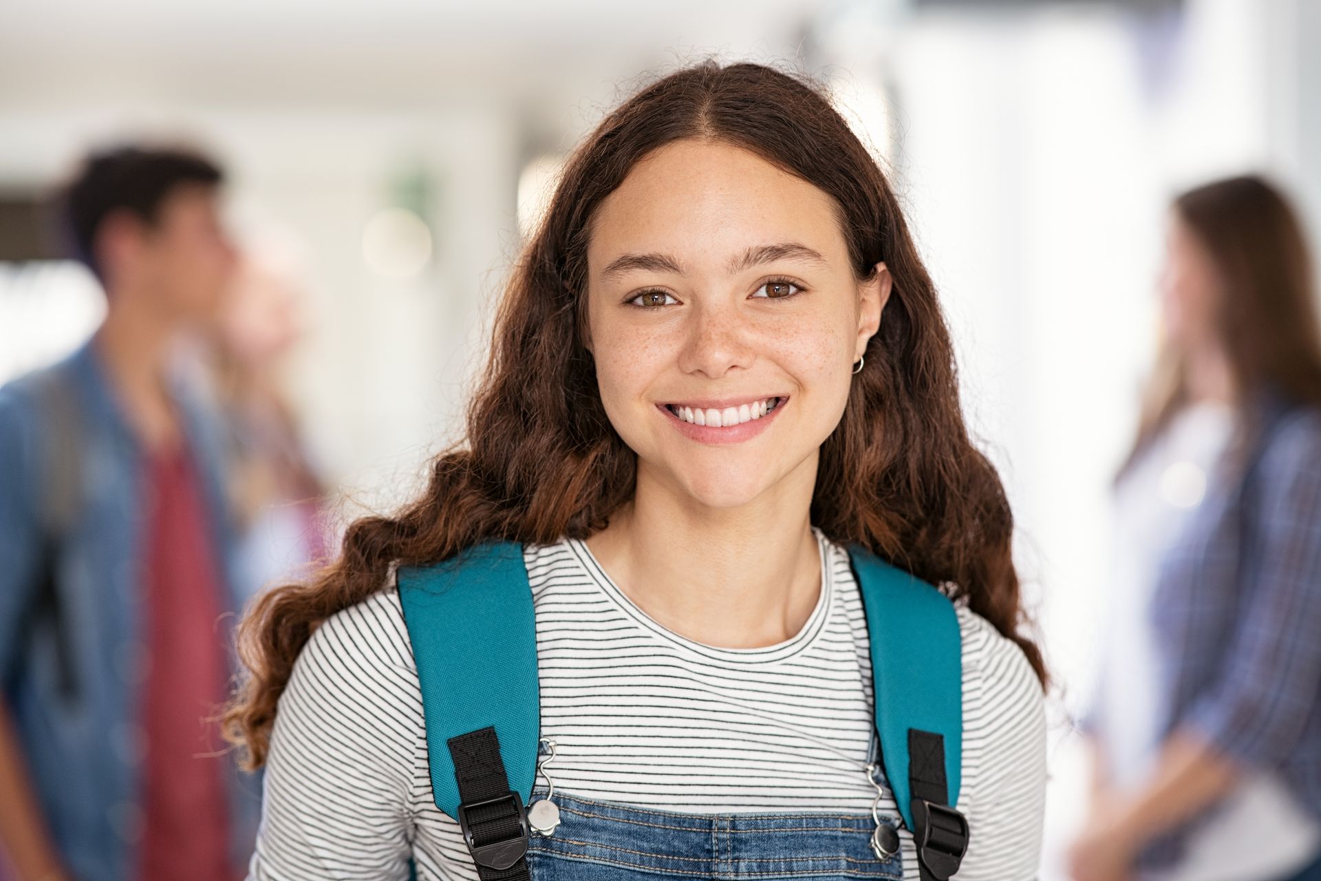 Smiling student with backpack in a school hallway, with classmates blurred in the background