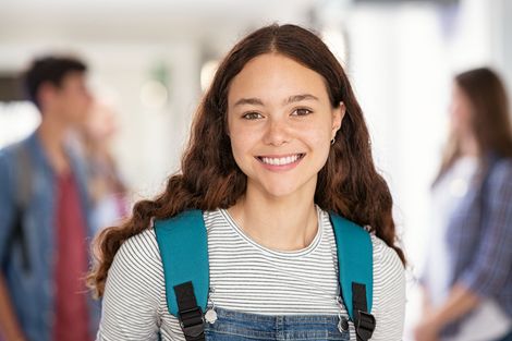 Smiling student with backpack in a school hallway, with classmates blurred in the background
