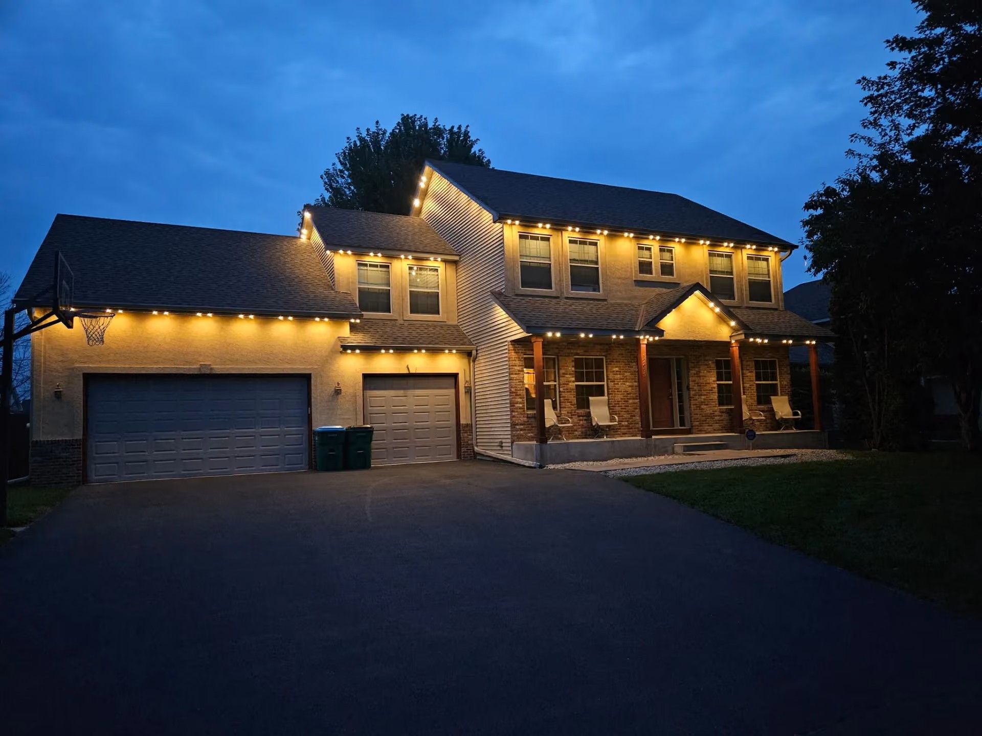 Large two-story house with warm exterior lights at dusk, featuring a garage and front porch.