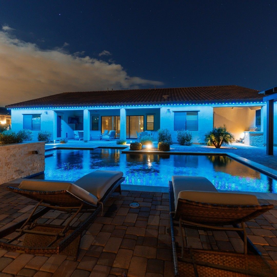 Blue-lit backyard pool at dusk beside a white house, with lounge chairs and glowing patio lights.