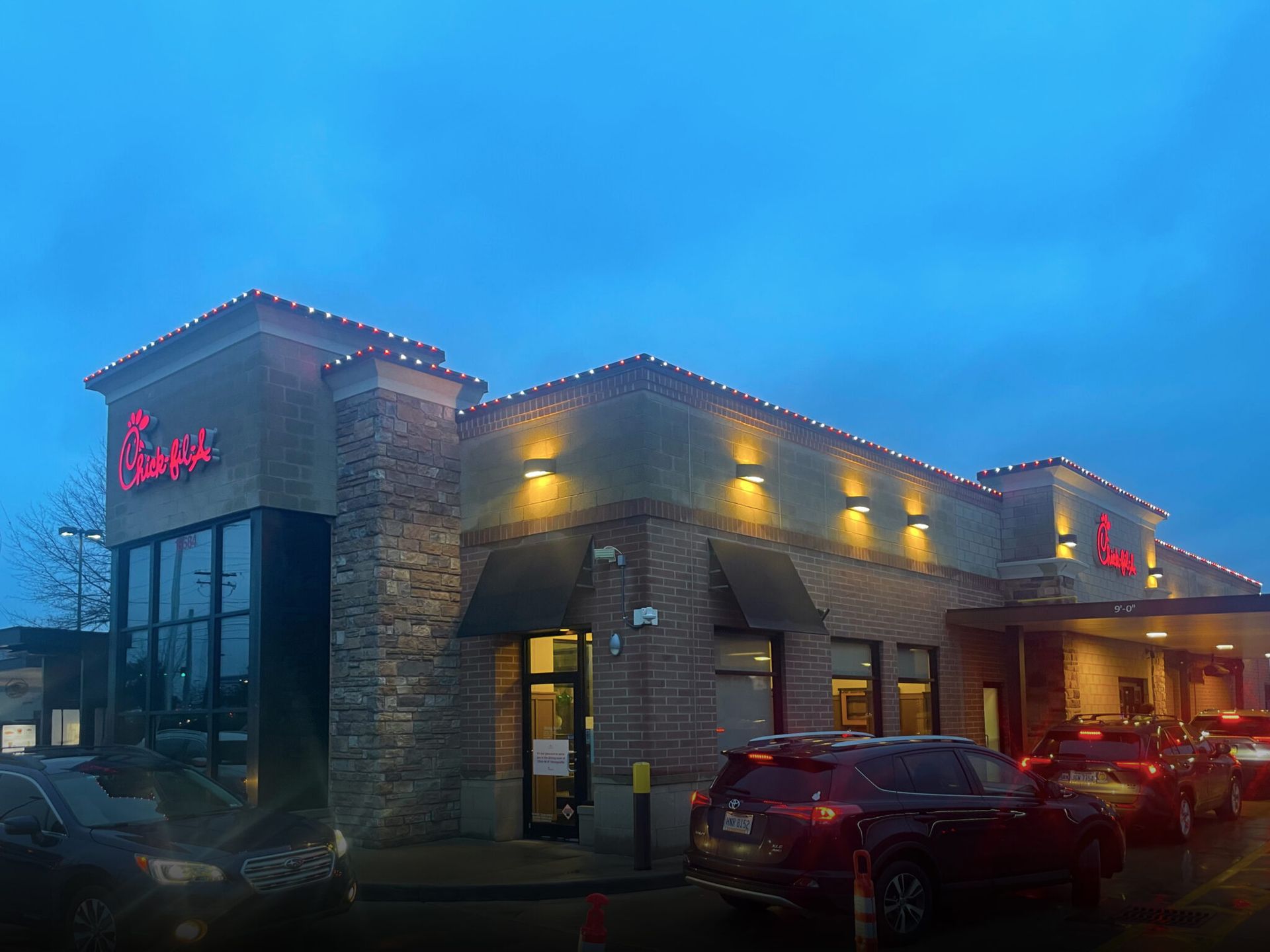 Brick restaurant at dusk with glowing lights, red sign, and cars parked outside.