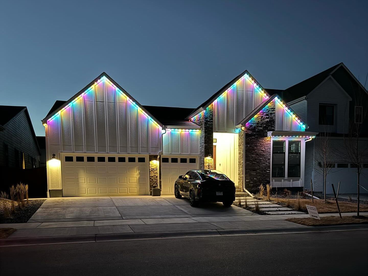 Suburban house decorated with multicolored Christmas lights at dusk, with a car in the driveway
