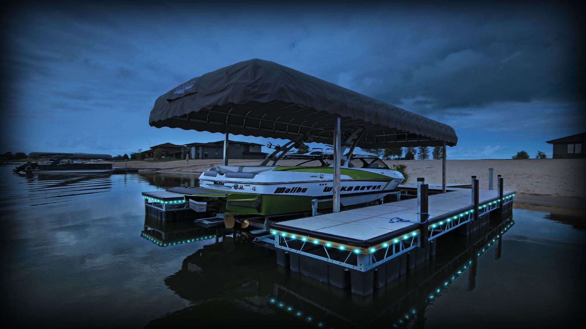 Covered dock with boats at dusk on a calm lake, lit by blue accent lights beneath a dark sky