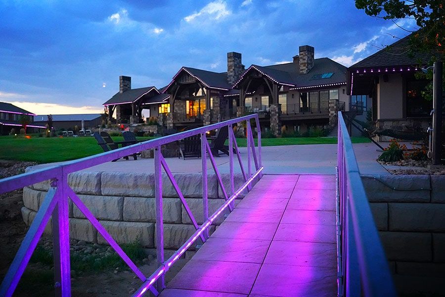 Purple-lit bridge leading to a lakeside lodge at dusk under a blue sky