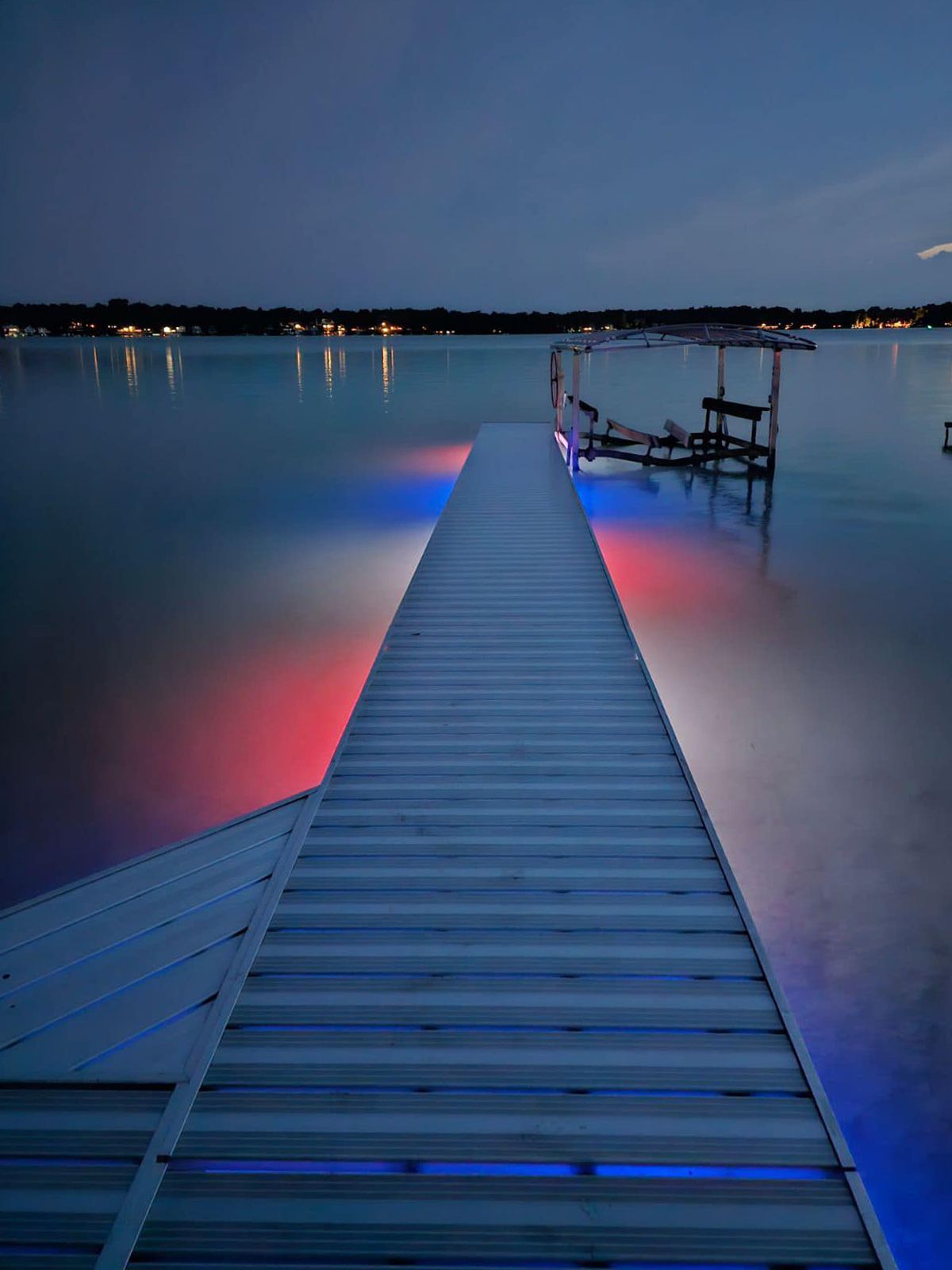 Illuminated dock extending into calm lake at dusk, with red and blue lights reflecting on the water