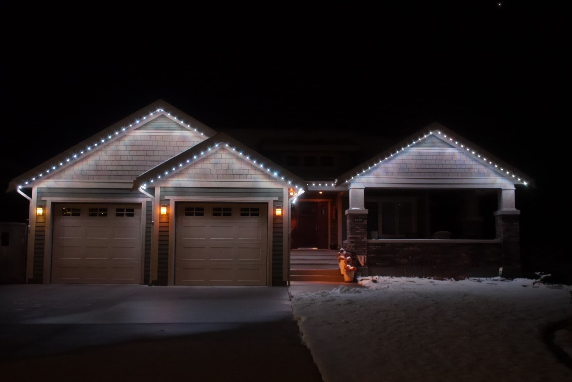 Snowy suburban house at night with Christmas lights outlining the roof and garage doors