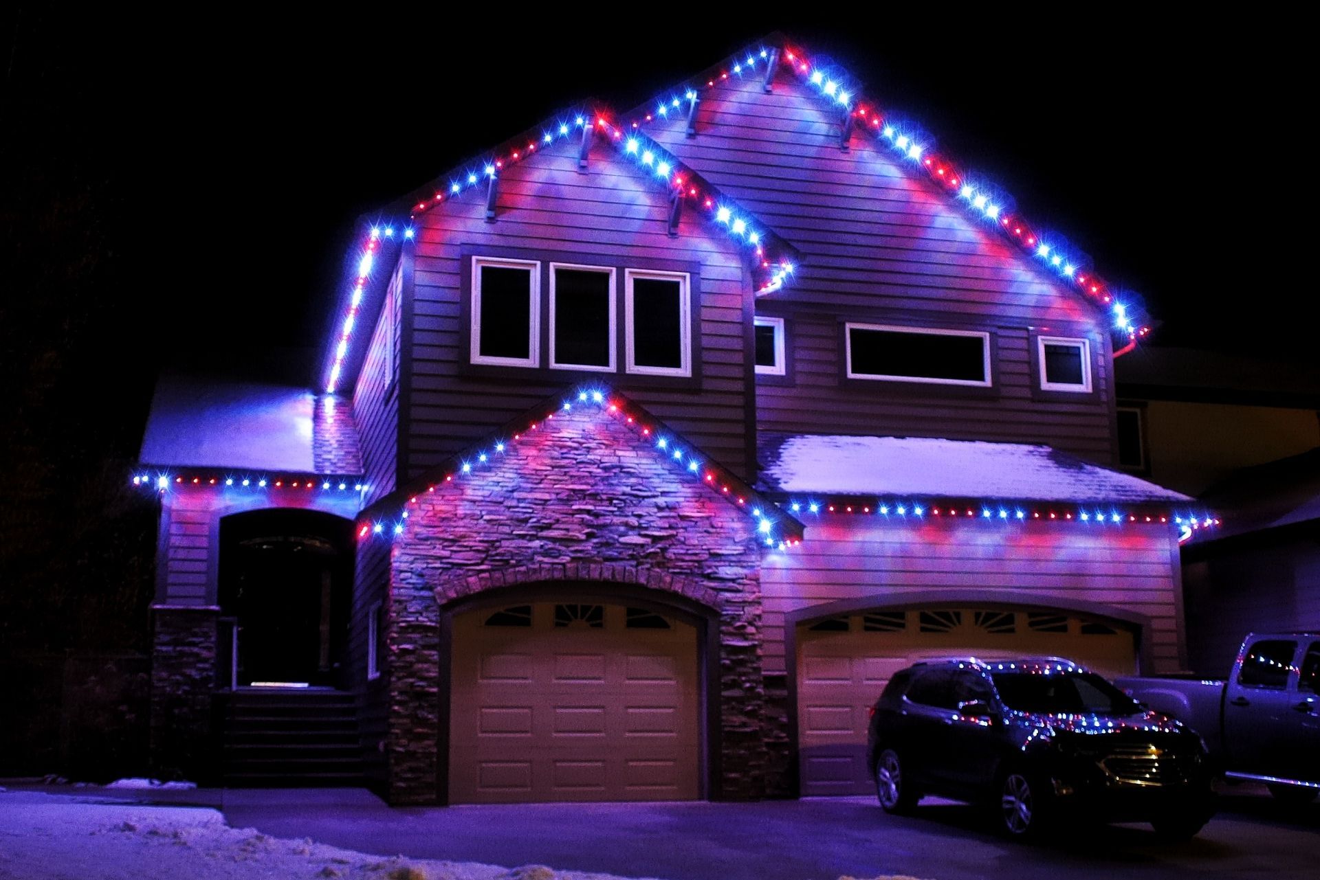 House decorated with red, blue, and white holiday lights at night, with cars parked in front.