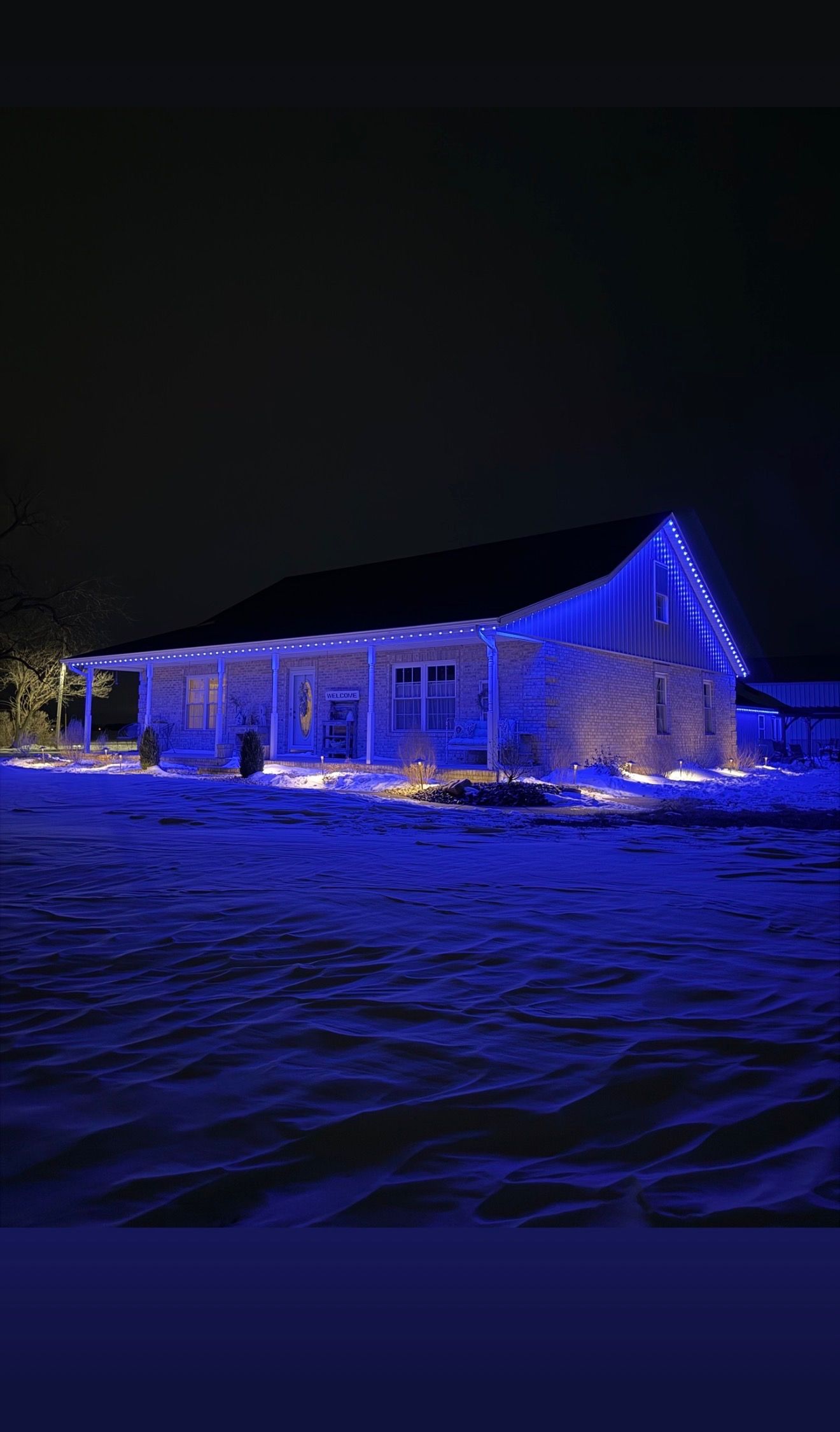 Snow-covered house lit in blue at night, with festive lights glowing against a dark sky