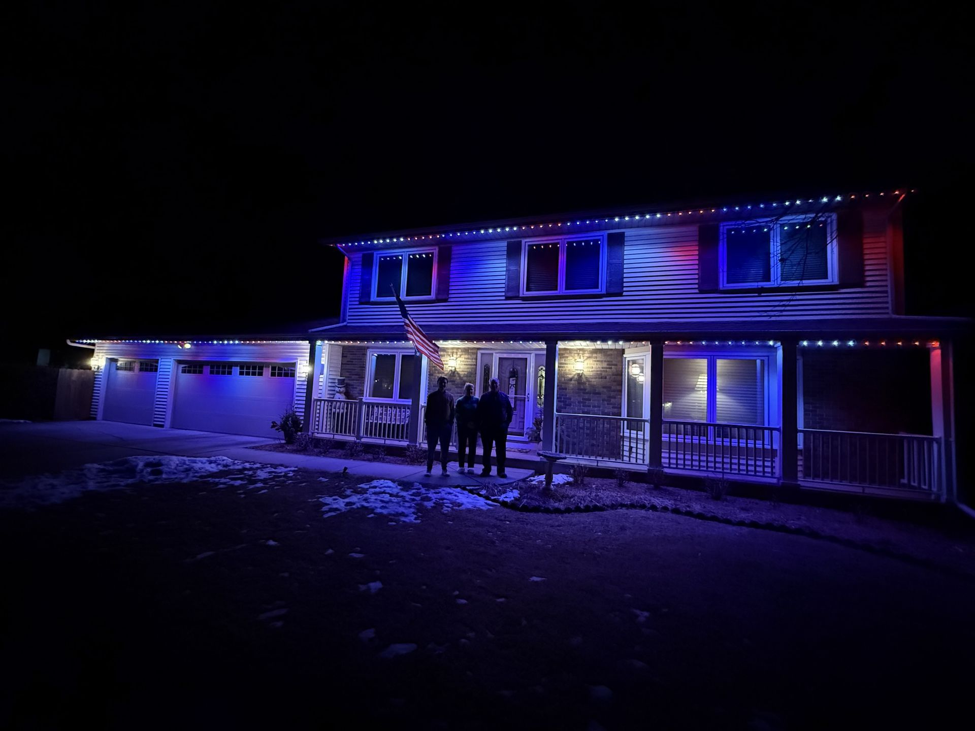House lit in blue and red at night, with people standing on the front porch and snowy ground.