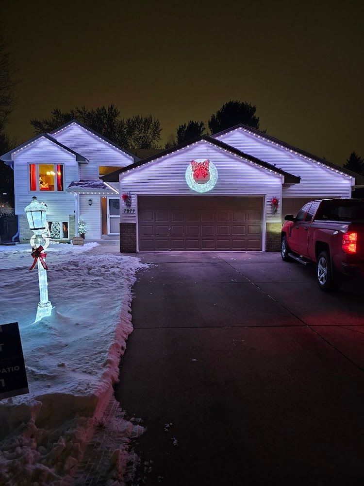 Snowy suburban house at night with Christmas lights, wreaths, and a decorated driveway.