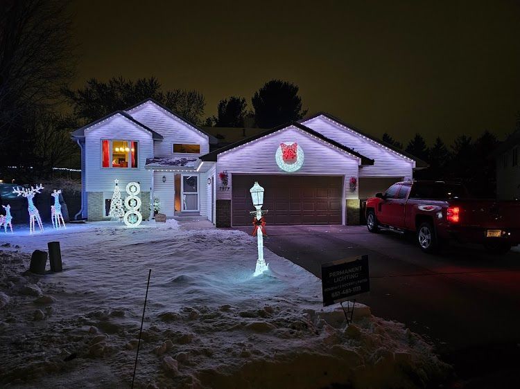 Snow-covered suburban house at night with colorful Christmas lights, glowing decorations, and a red pickup in the driveway