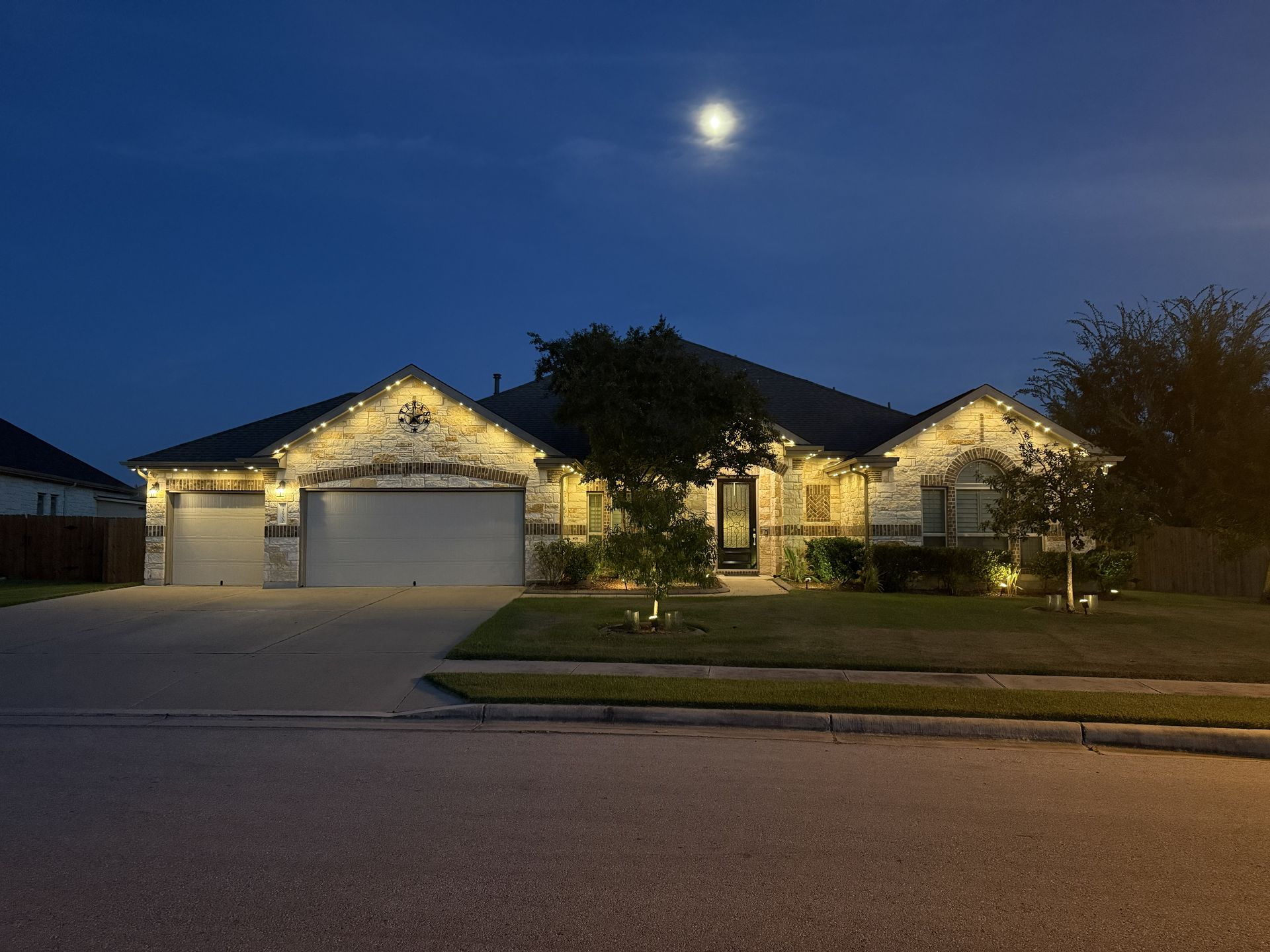 Suburban house at dusk with lit exterior lights and a bright moon overhead