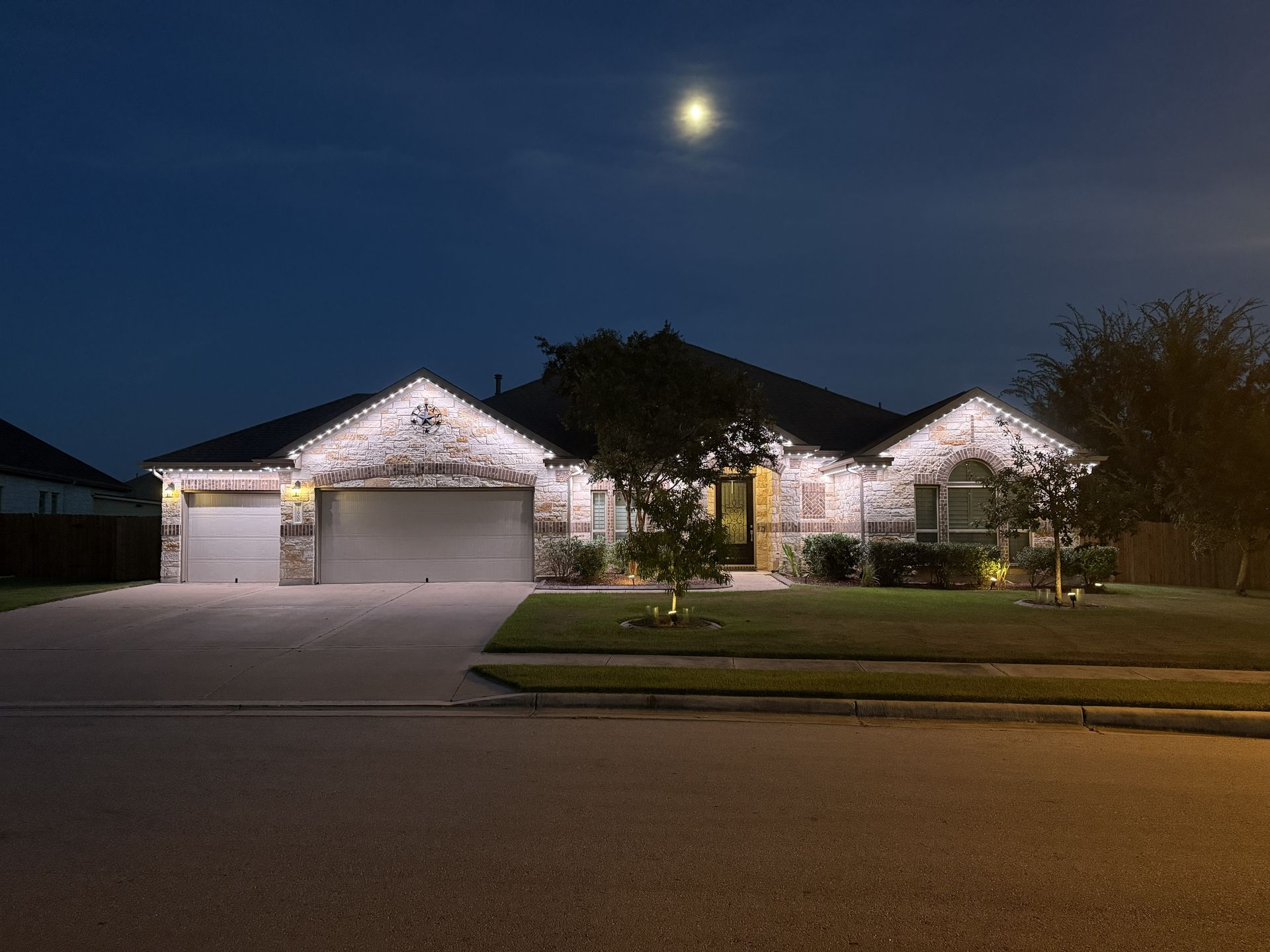 Single-story house at night with lights on, two-car garage, and a bright moon overhead