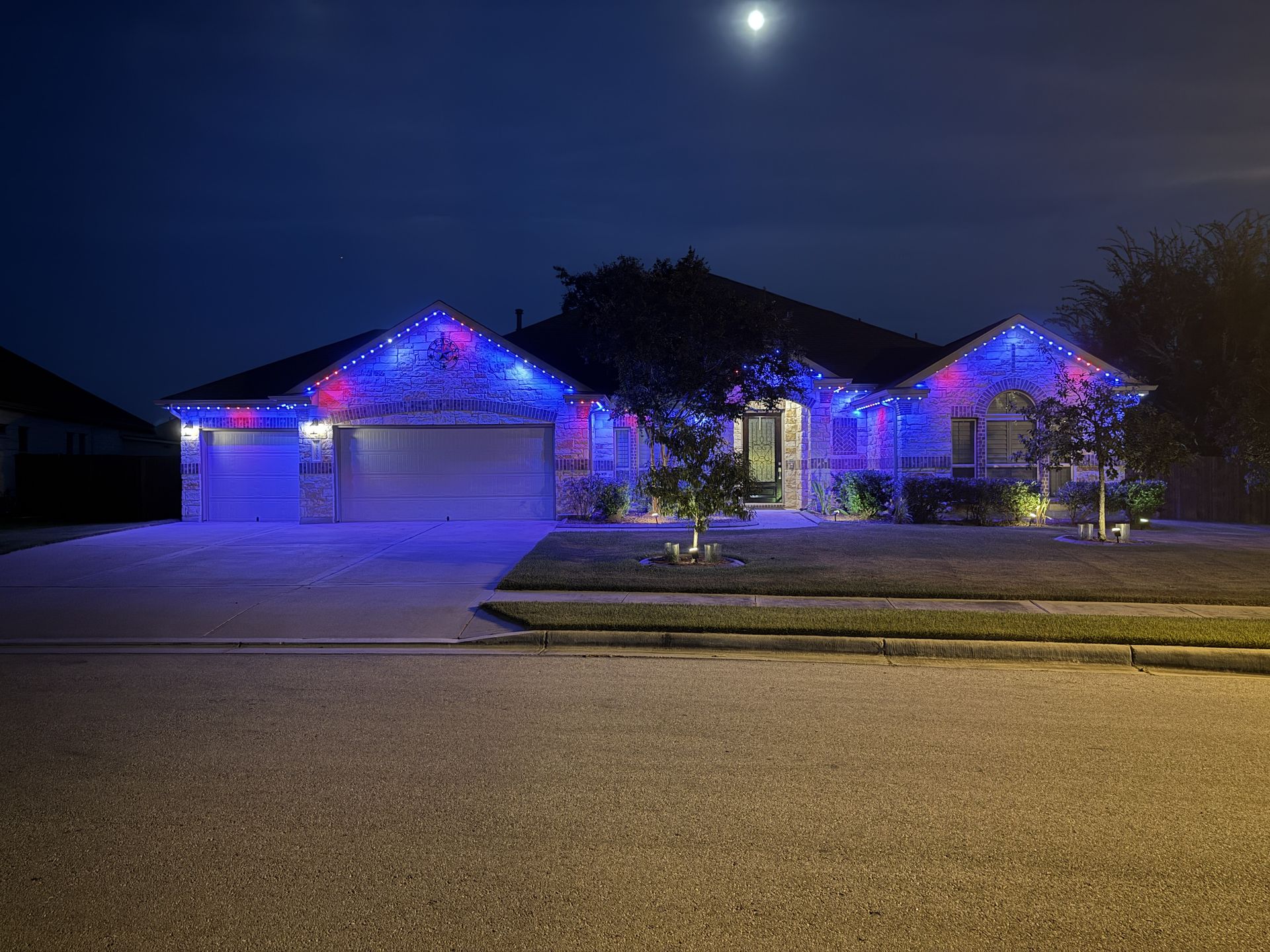 Single-story house at night with blue and red Christmas lights, lit under a bright moon.