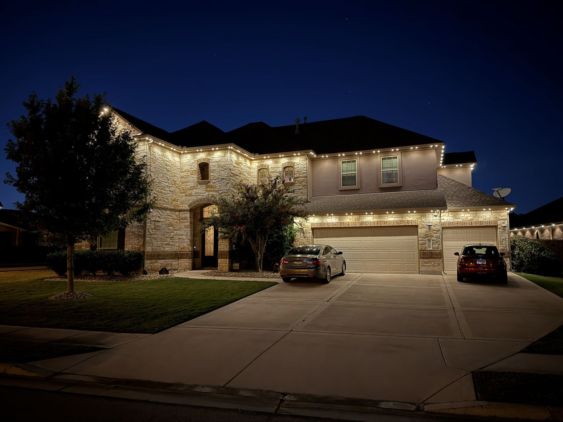 Two-story house at dusk with warm lights, decorated roofline, and cars parked in the driveway