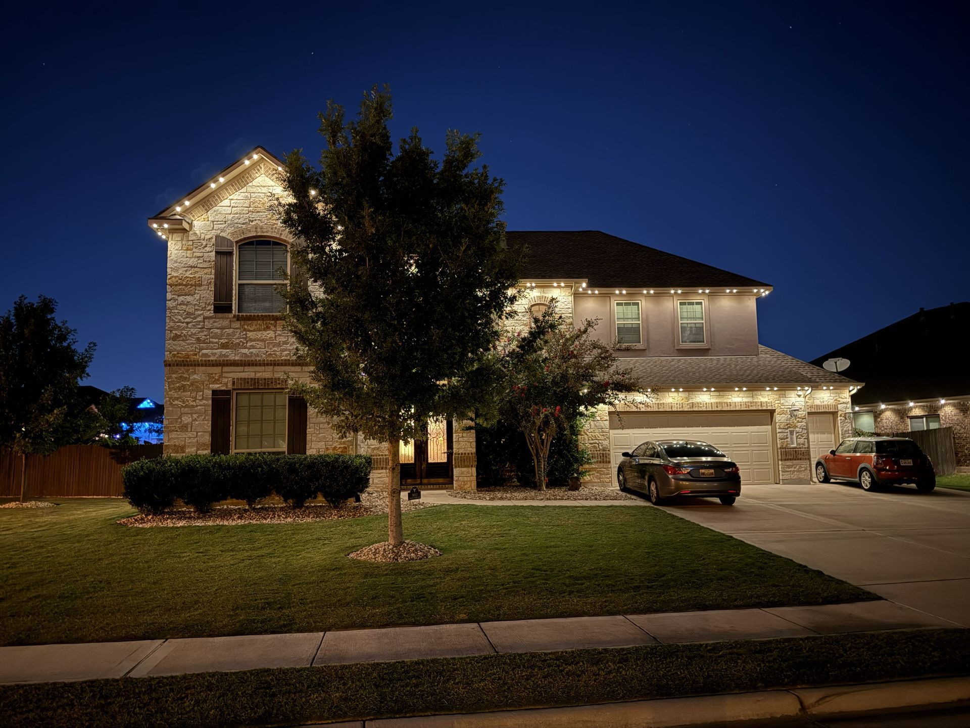 Two-story suburban house at night with lights on, a tree in front, and cars parked in the driveway
