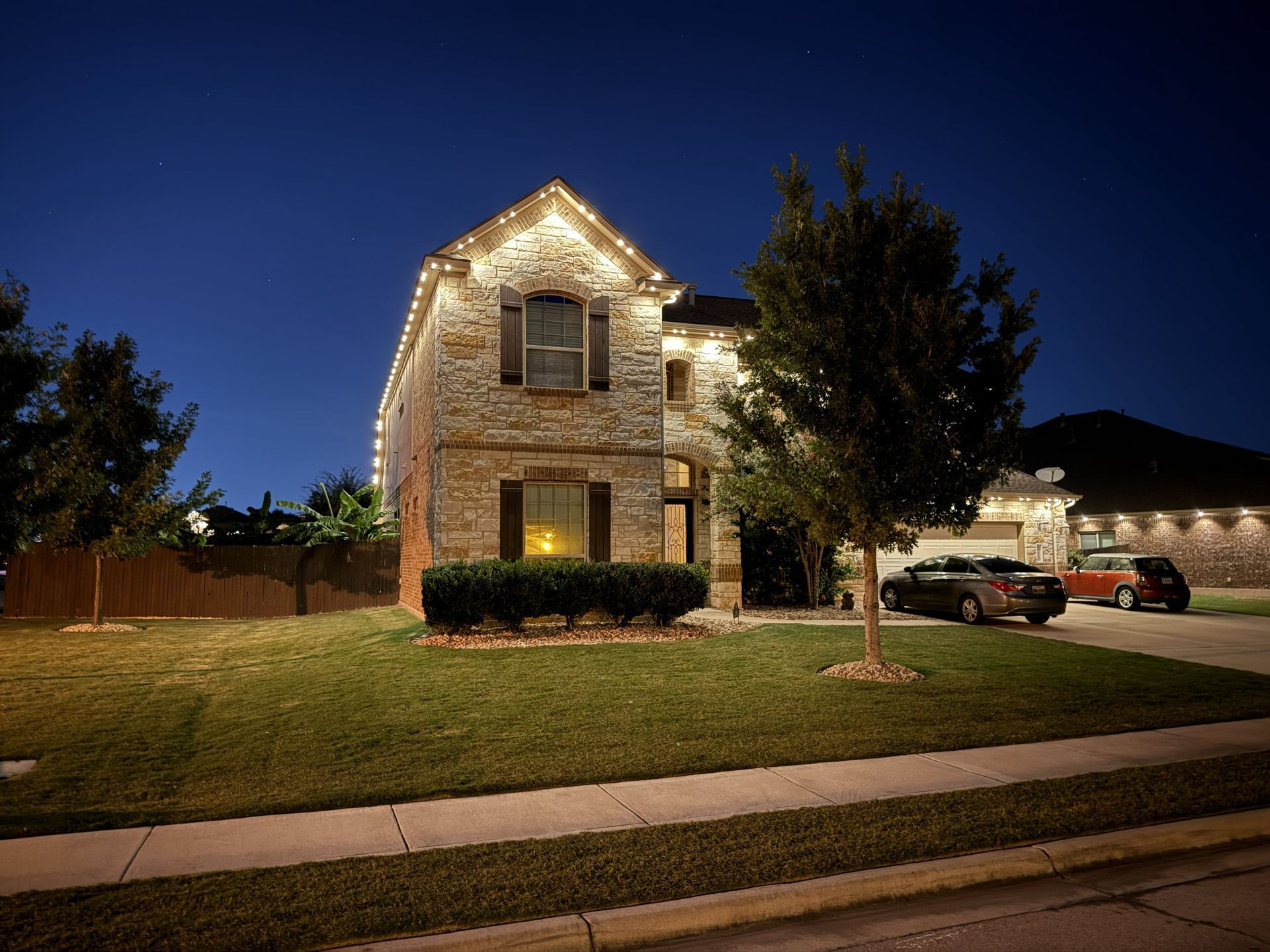 Two-story brick house lit with white lights at night, with a lawn, driveway, and parked cars.
