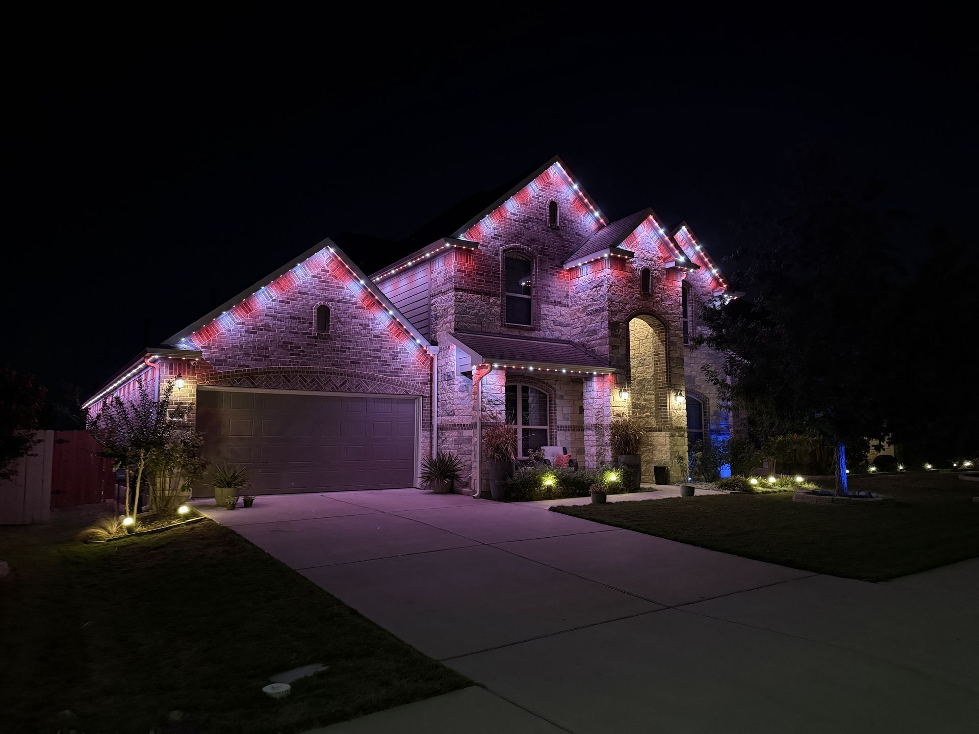 Suburban house at night lit with pink and purple holiday lights, with a driveway and dark lawn