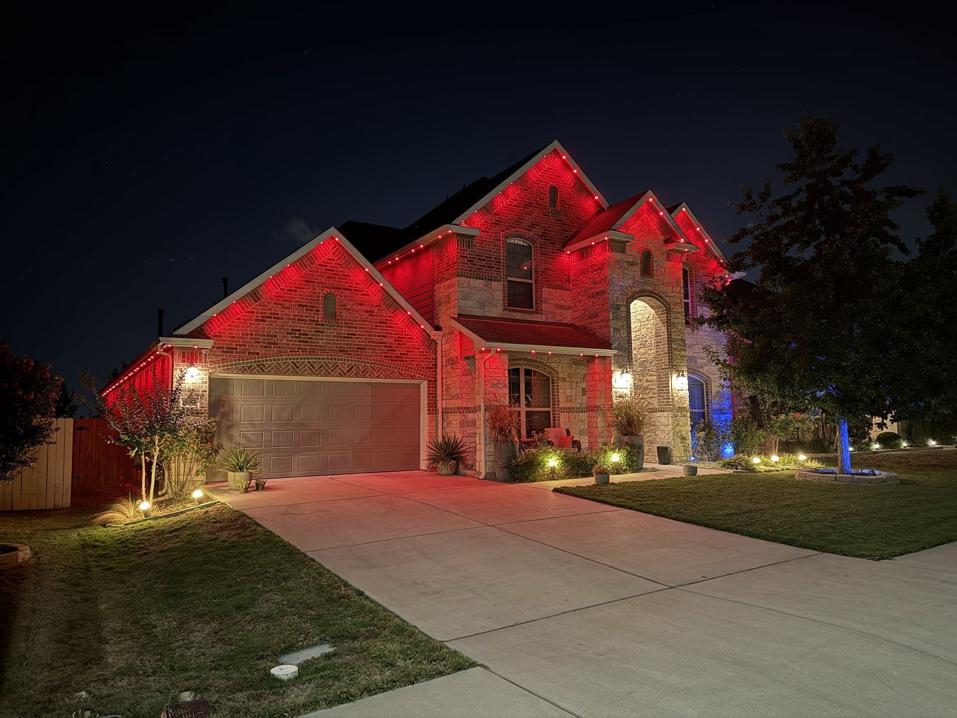 Two-story suburban house lit with red holiday lights at night, with a driveway and lawn