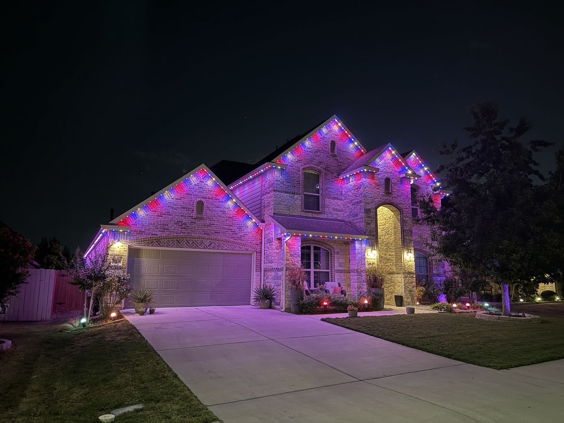 Suburban house at night lit with purple and pink holiday lights, with a driveway and lawn visible