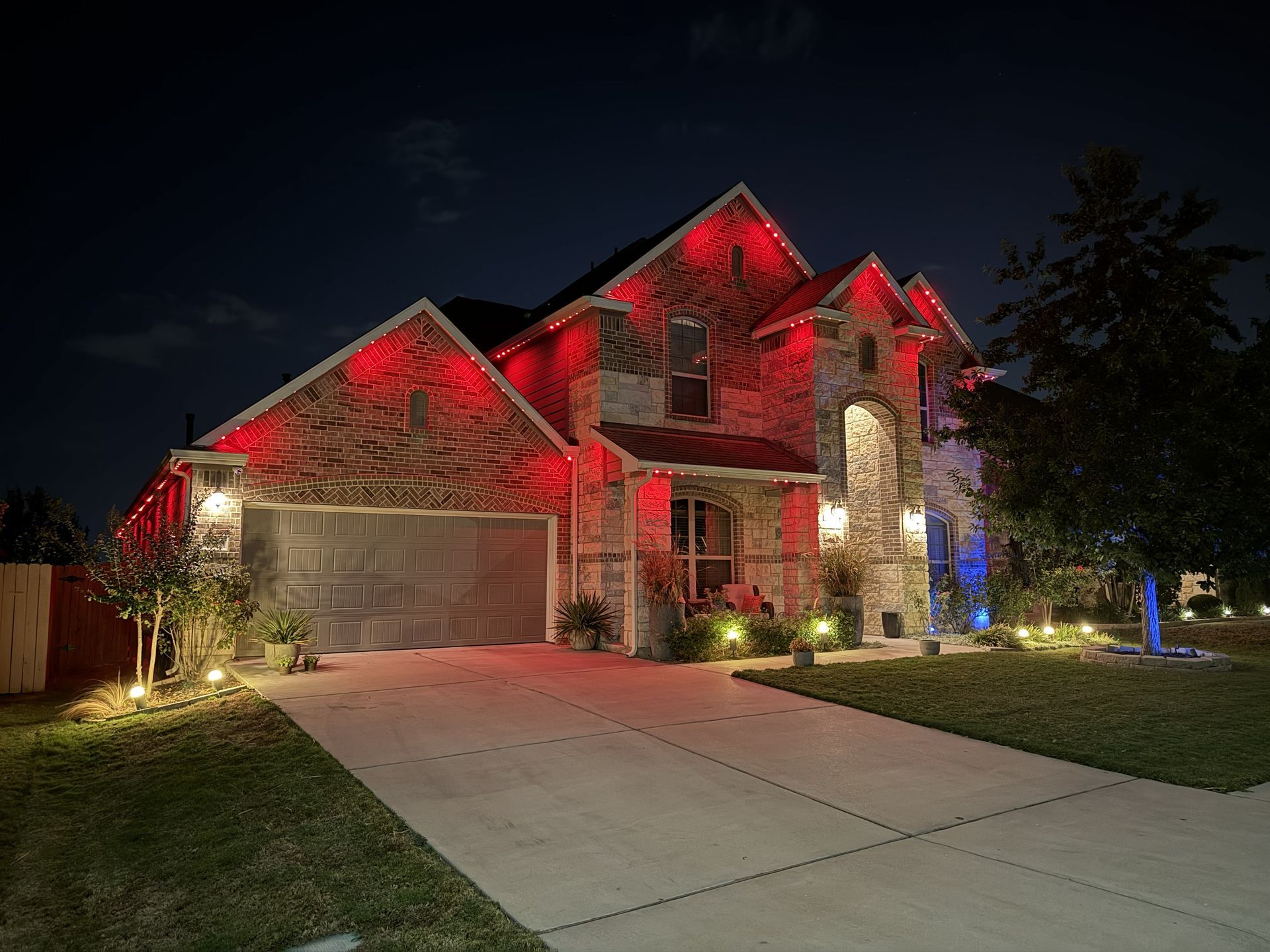 Suburban house at night lit with red and white holiday lights, with a driveway and lawn in front