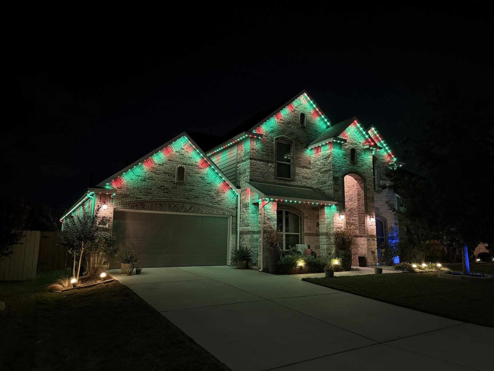 Suburban house at night decorated with red and green Christmas lights, with a lit driveway and garage.