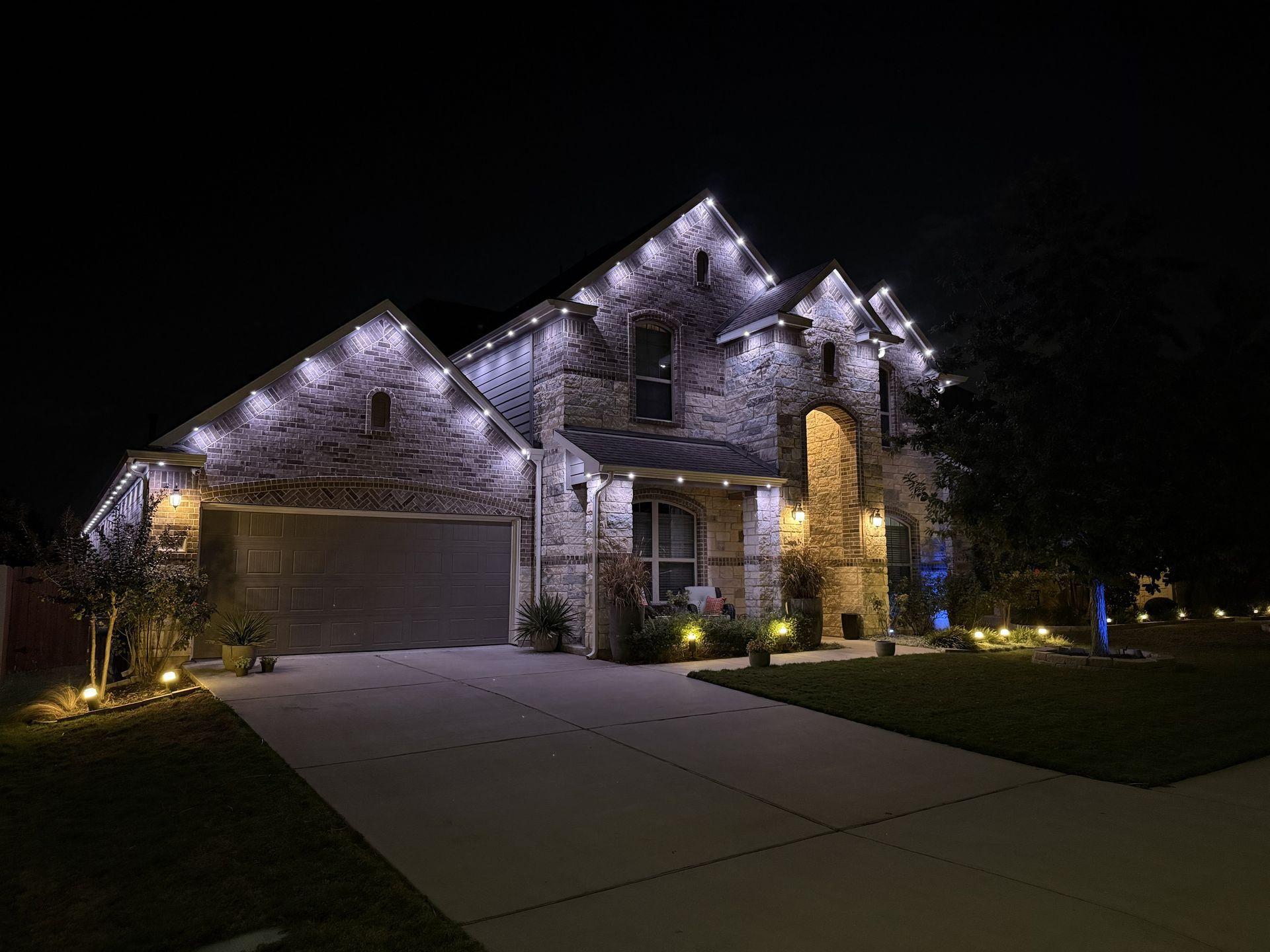 Two-story stone house lit with white lights at night, with a driveway and front lawn.