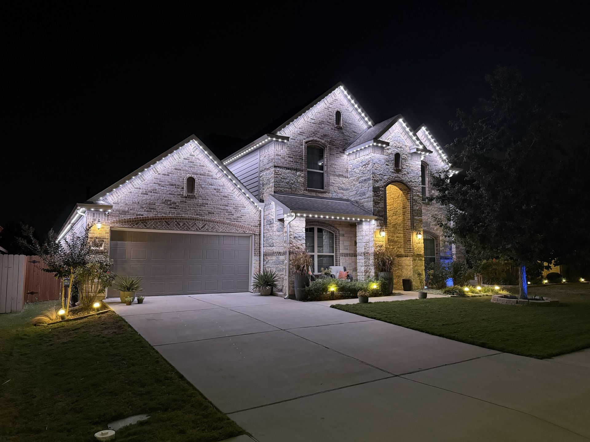 Illuminated stone house at night with a garage, driveway, and lit landscaping