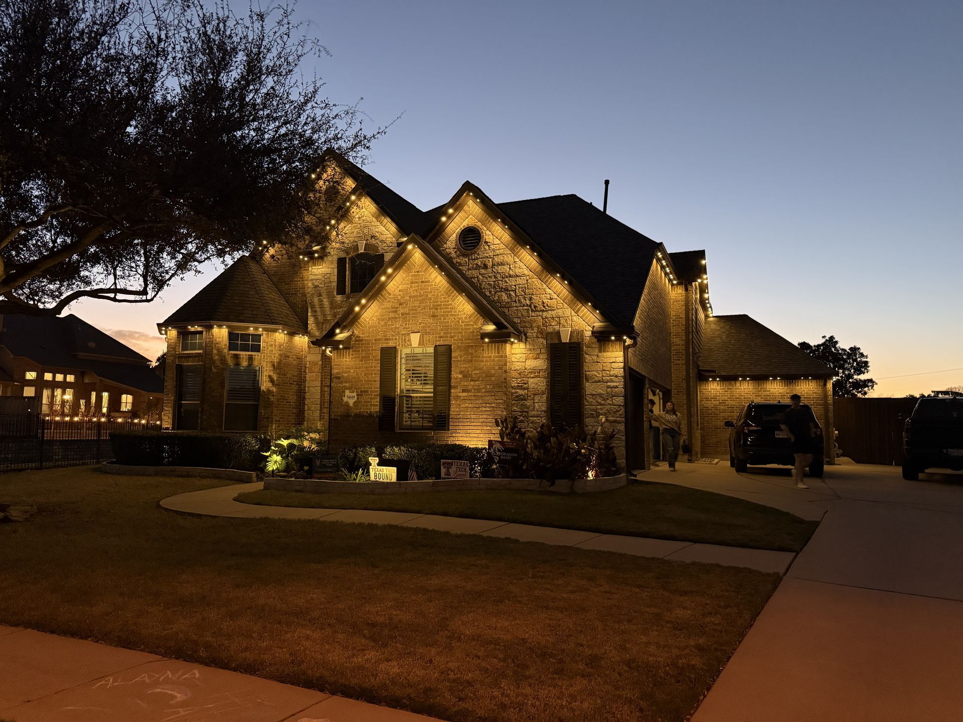 Brick house lit at dusk with warm porch lights, driveway, and a darkening sky