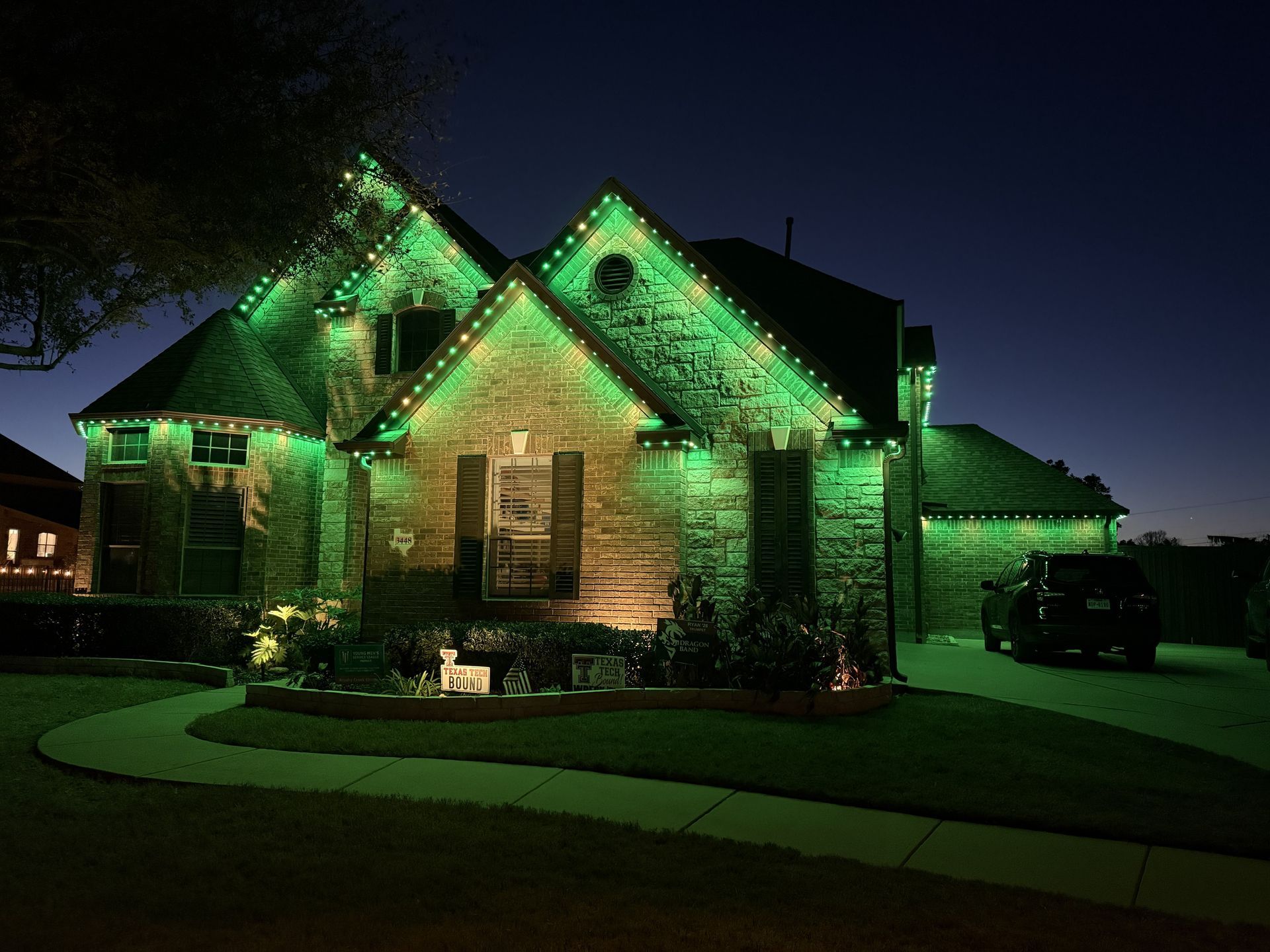 Brick house lit with green lights at night, with a curved driveway and dark sky.