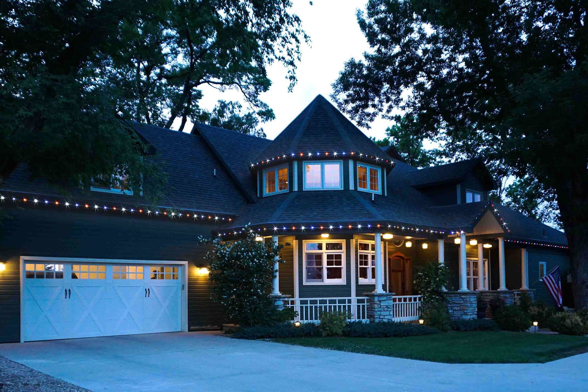 Suburban house at dusk with lit windows, white garage, and string lights along the roofline
