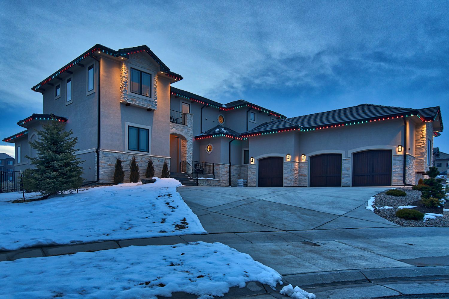 Snowy suburban house at dusk with warm lights and a garage driveway