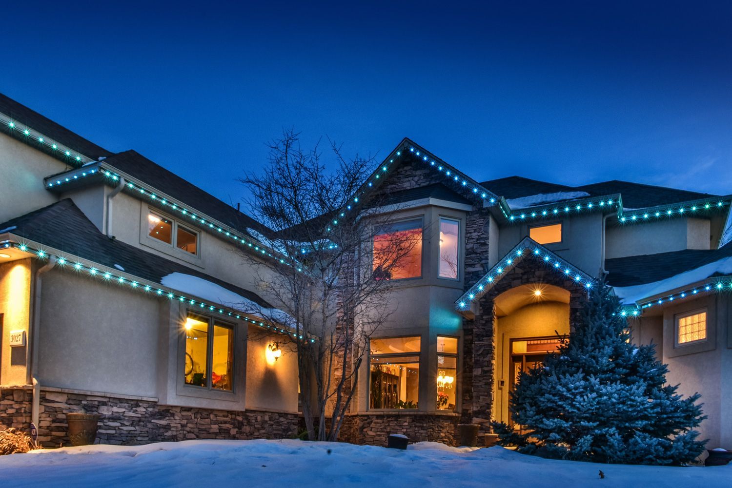 Snowy house at dusk with blue holiday lights glowing along the roofline and windows.