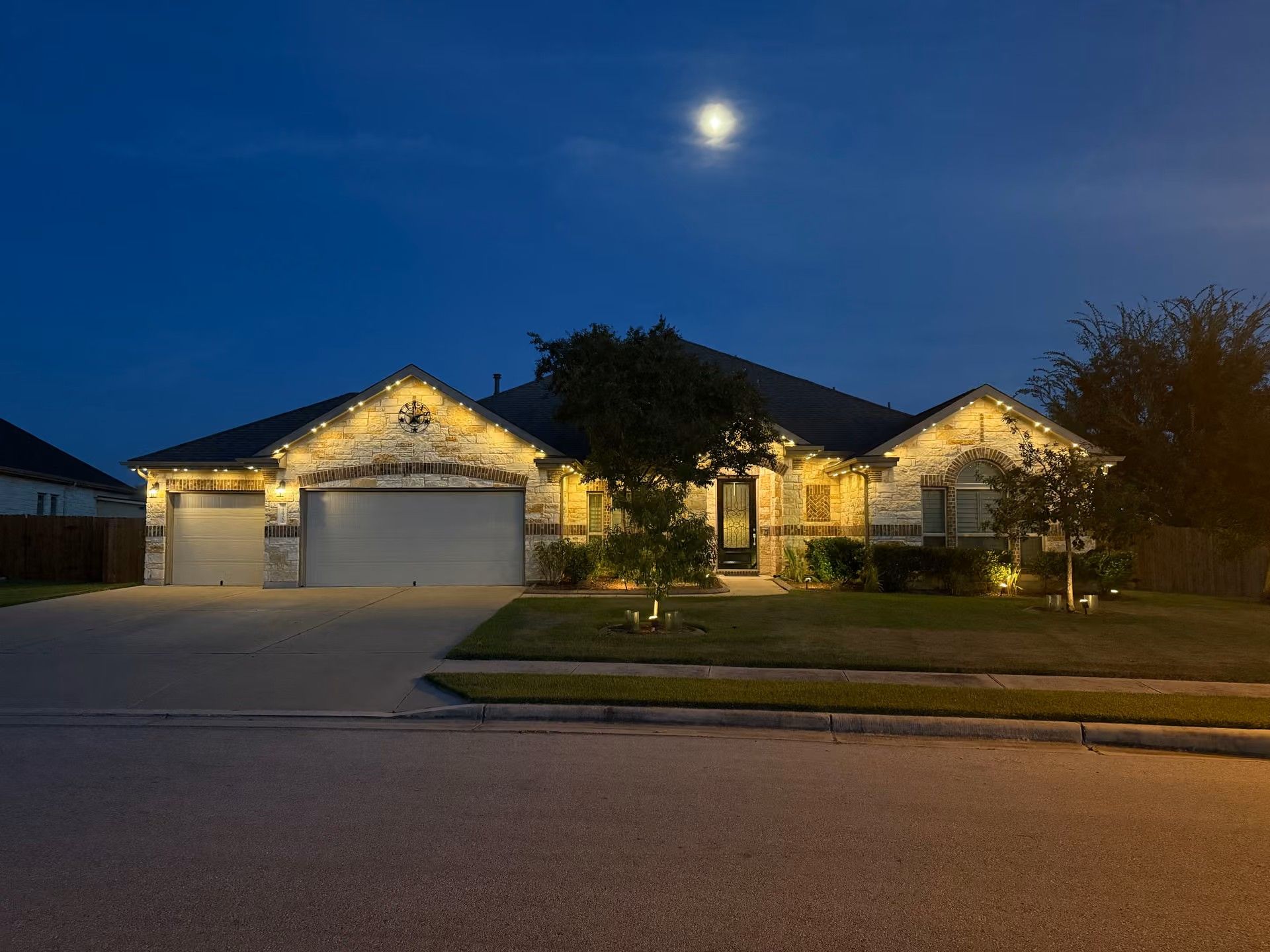 Suburban house at dusk with warm porch lights, two garages, and a bright moon in the sky