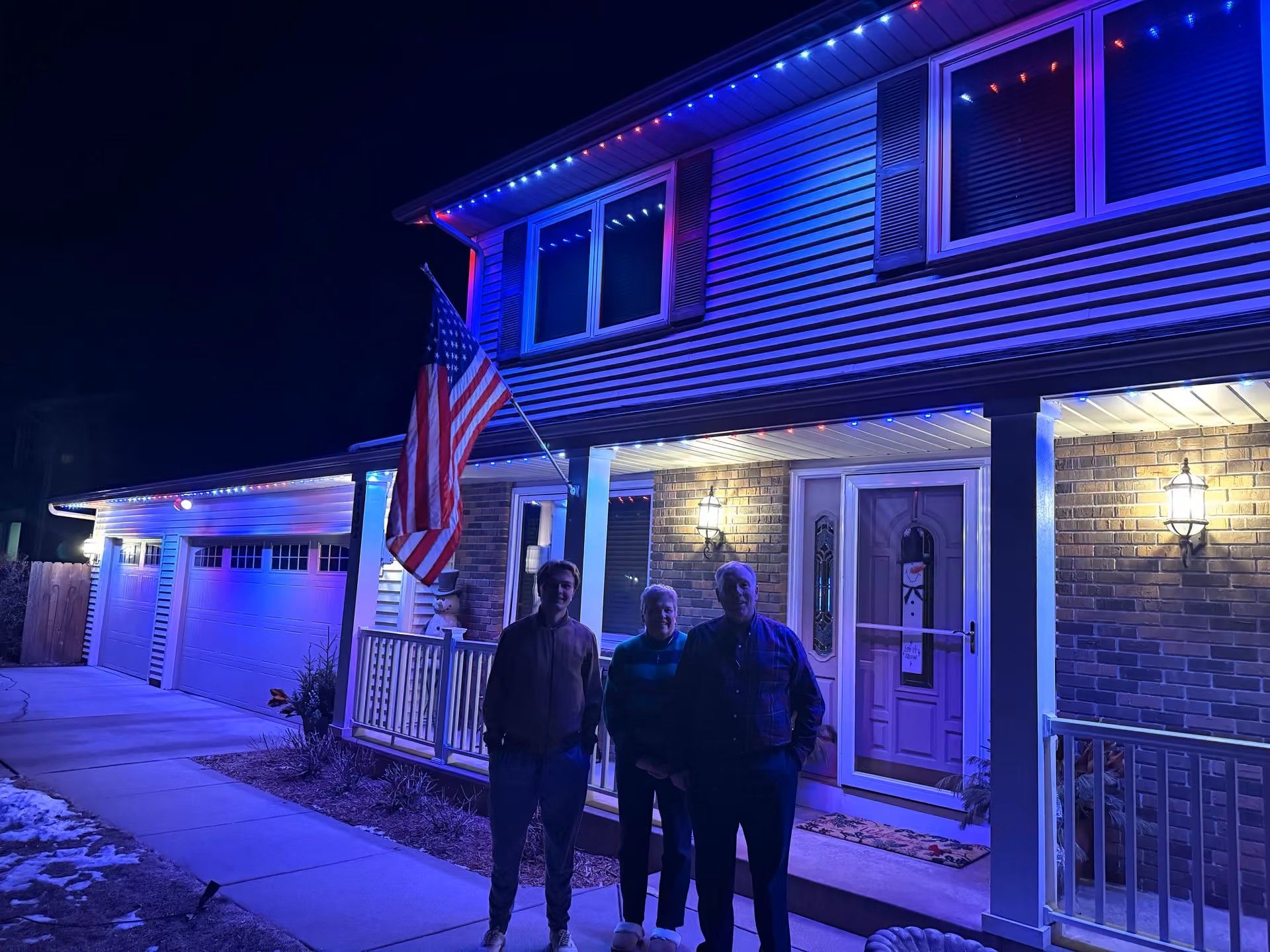 Two people standing outside a house at night, lit by blue and purple lights, with an American flag by the porch